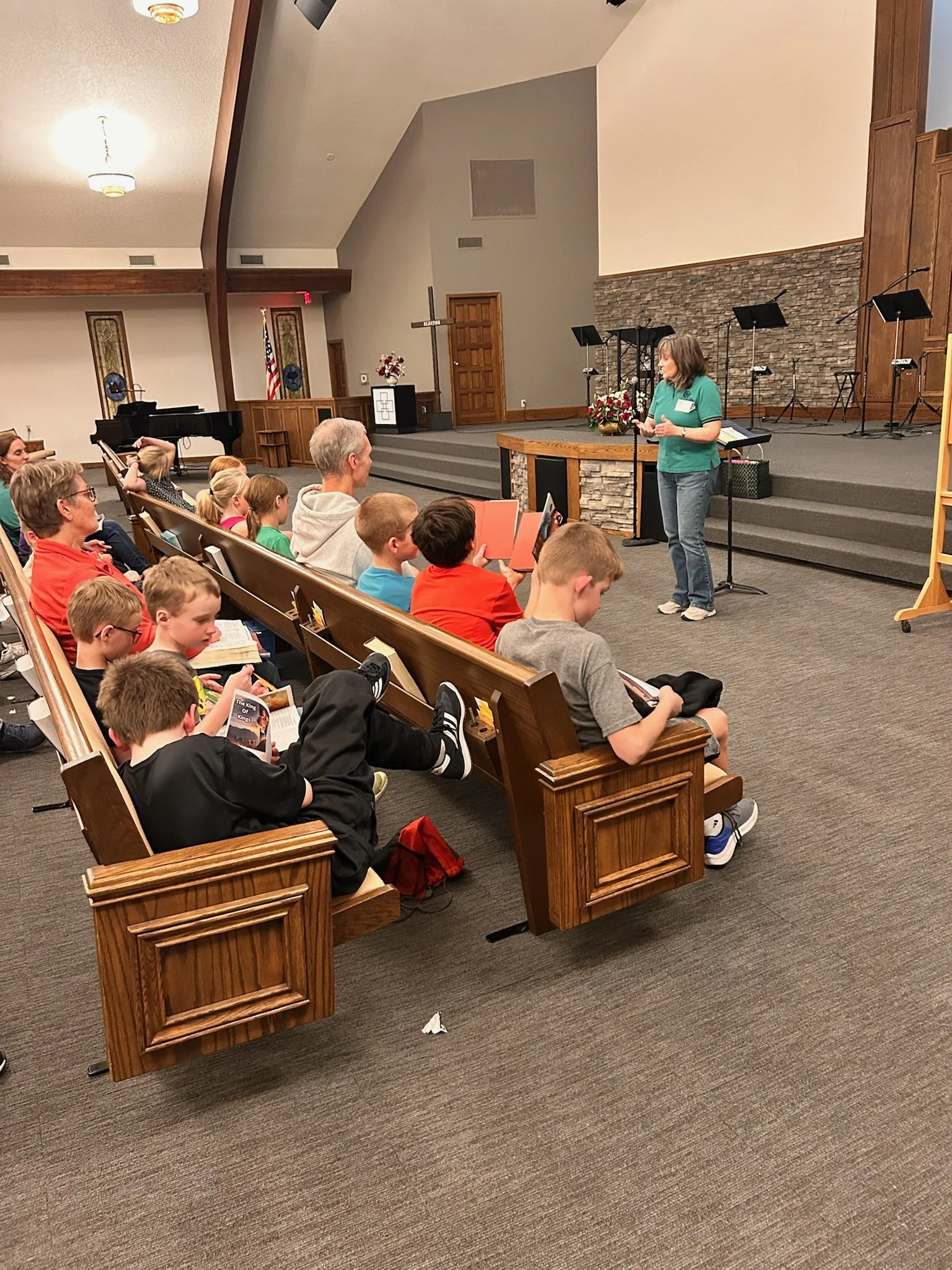 A woman speaking in front of a group of children seated in wooden pews during a church event. The stage behind her is decorated with flowers and has microphones, music stands, and a brick wall. The church has high ceilings, wooden accents, and stained-glass windows.