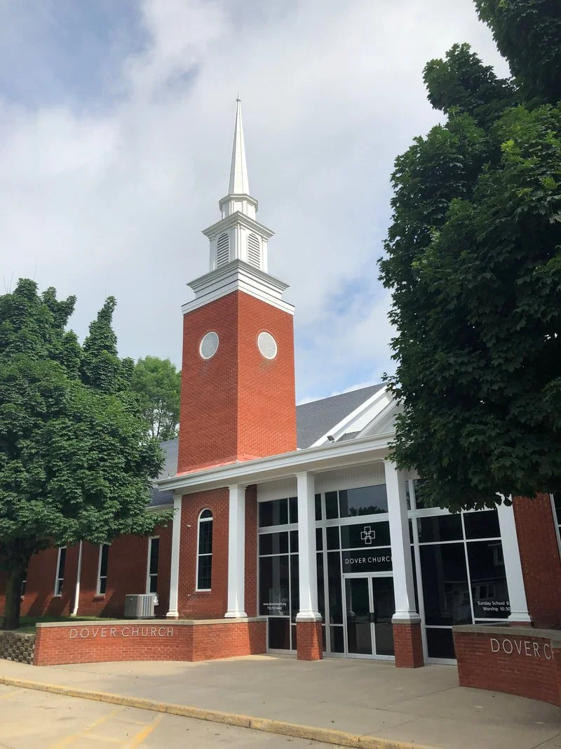 Exterior view of Dover Church, a brick building with a tall white steeple, large windows, and entryway with white columns, surrounded by green trees.