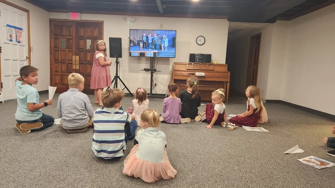 A group of children sitting on the carpeted floor watching a girl in a pink dress singing or speaking into a microphone in front of a screen.