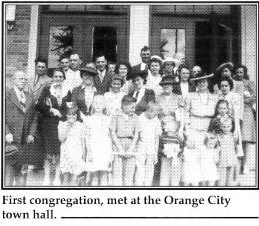 A group of people, including men, women, and children, standing in front of a building, posing for a group photo at the Orange City town hall.