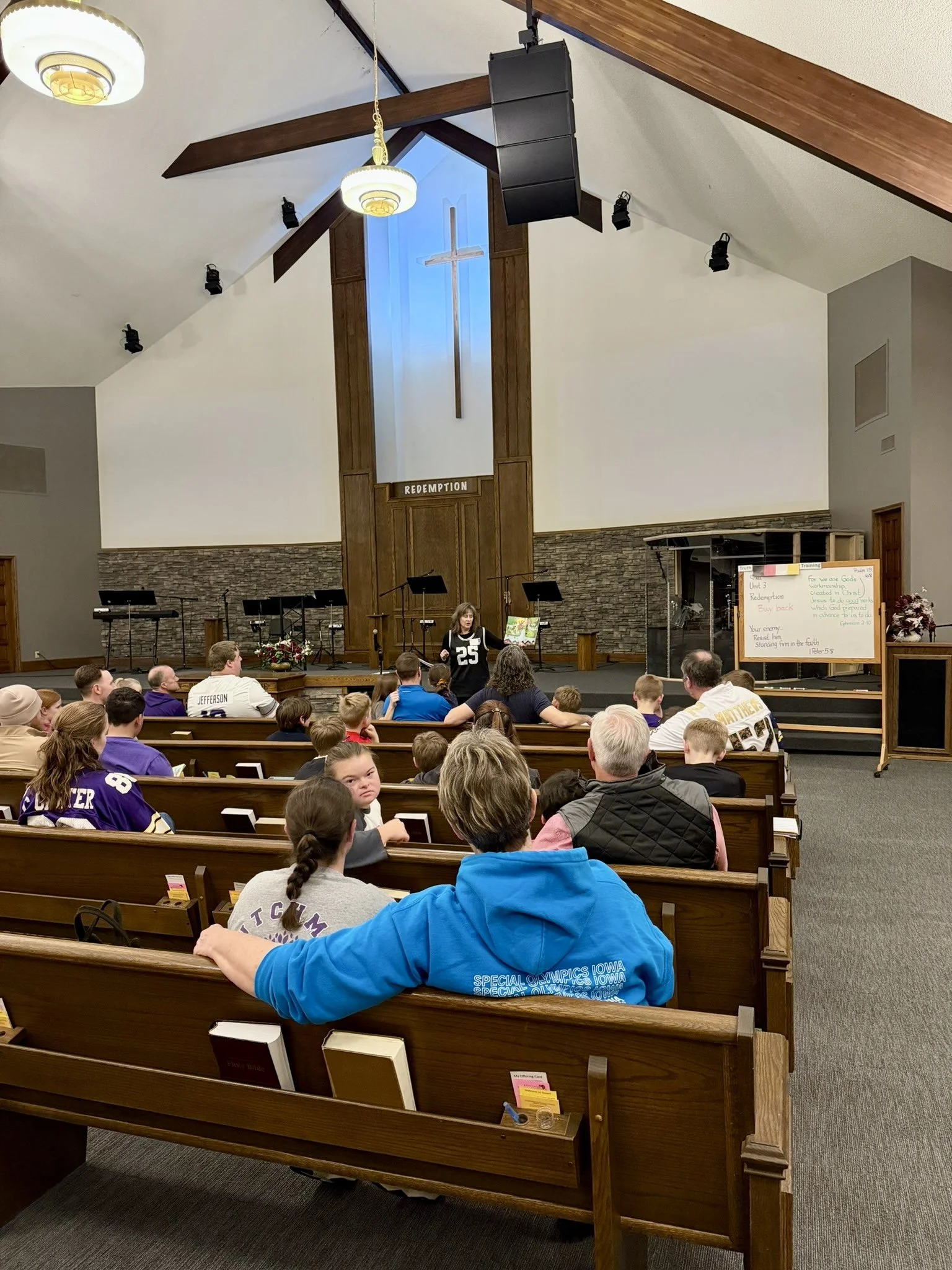 People seated on wooden pews inside a church, with a woman speaking at a podium in front of a cross and a sign that reads 'REDEMPTION'. The church interior features a high ceiling with wooden beams, a large blue window behind the cross, and multiple black speakers and lights.