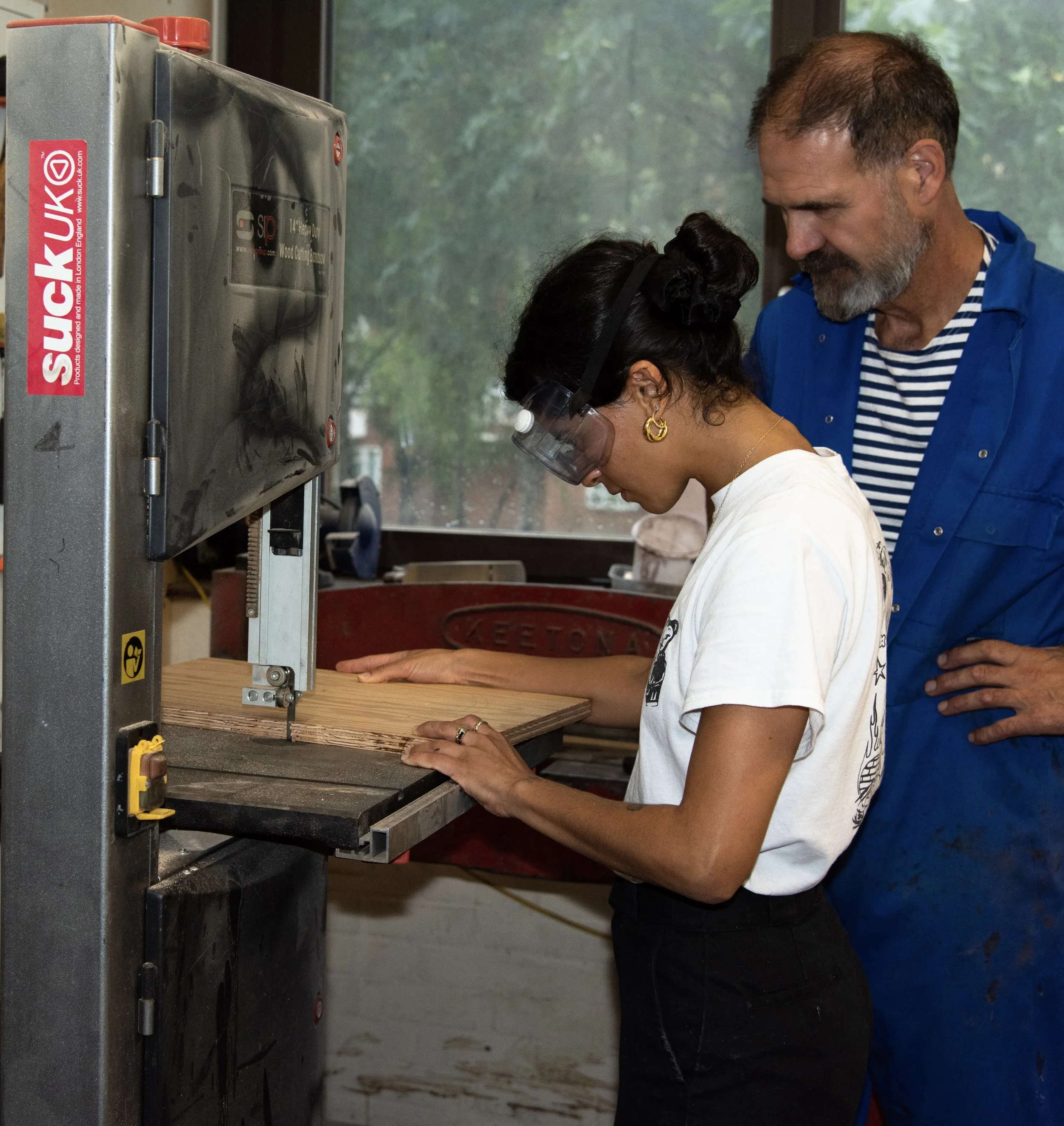 A woman operating a woodworking machine, possibly a planer or jointer, with a man standing behind her.