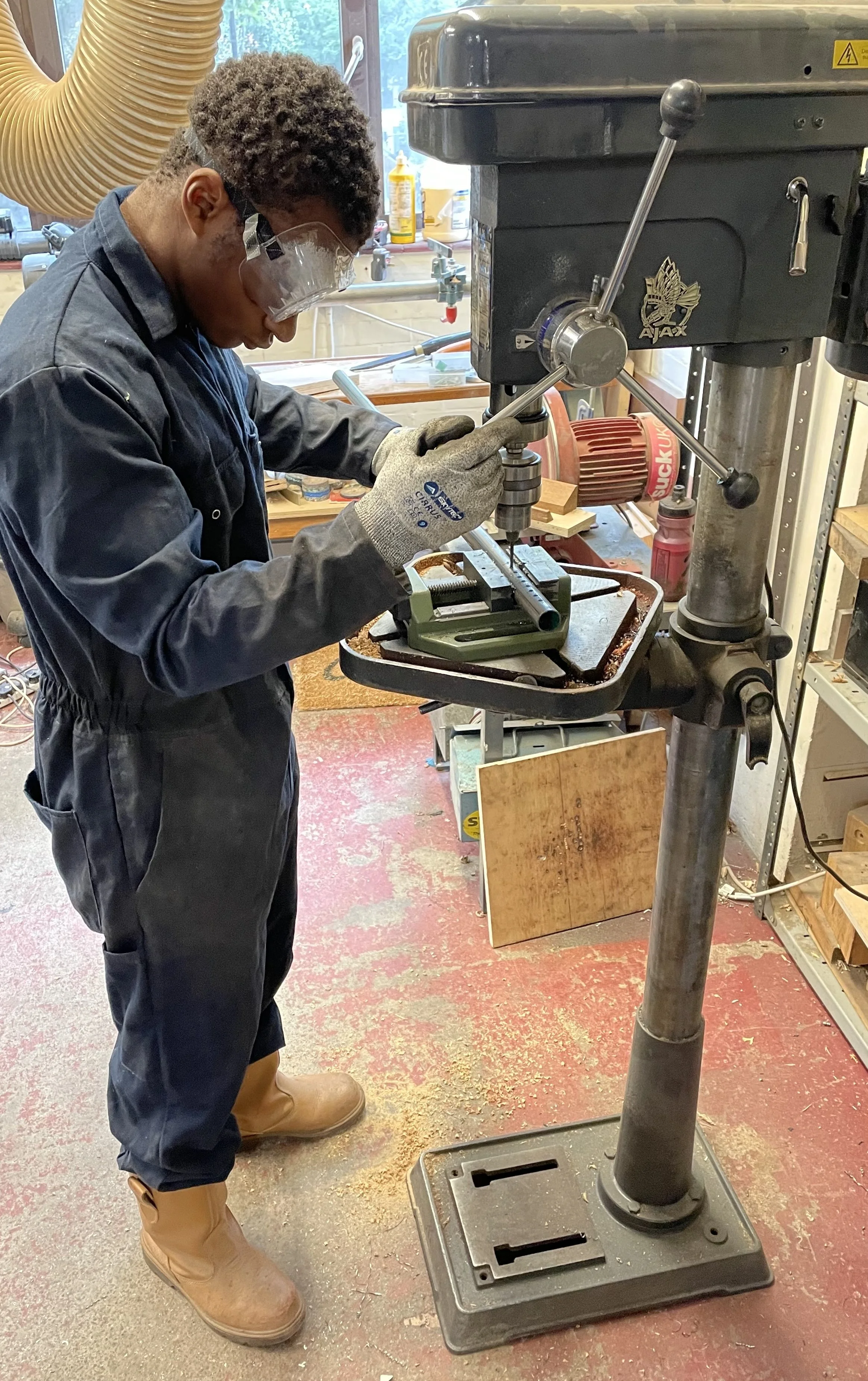 A person in work overalls and safety gear using a drill press in a workshop. The workspace includes various tools, safety supplies, and wood pieces.