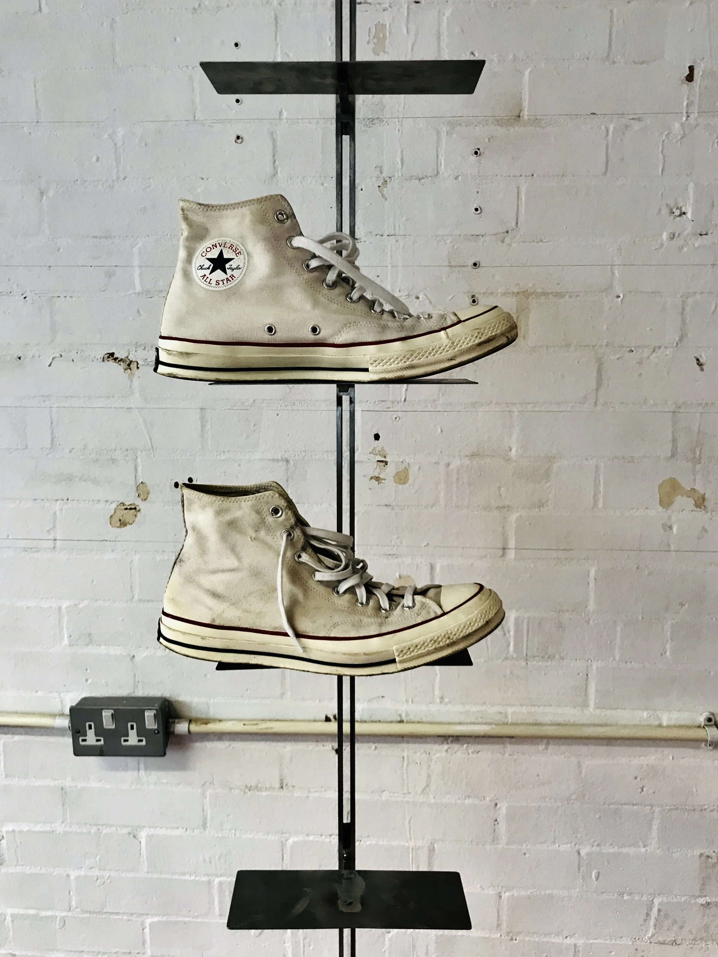 A pair of worn beige high-top Converse sneakers displayed on a black metal shelf mounted on a white brick wall.
