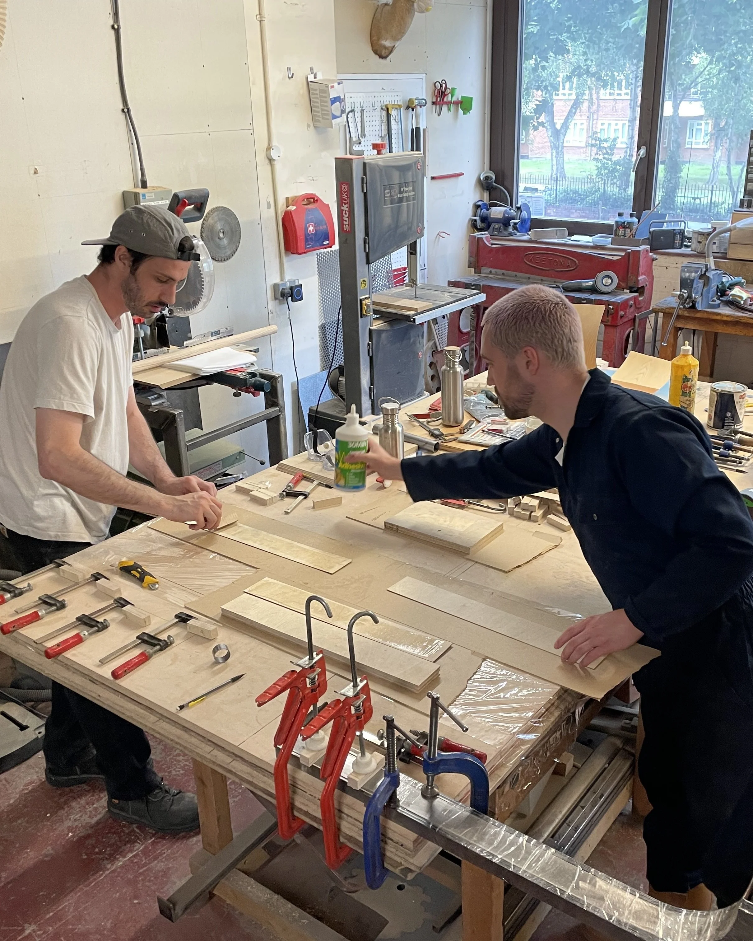 Two men working in a woodworking workshop, surrounded by woodworking tools, clamps, and pieces of wood, with a large window showing outside trees.