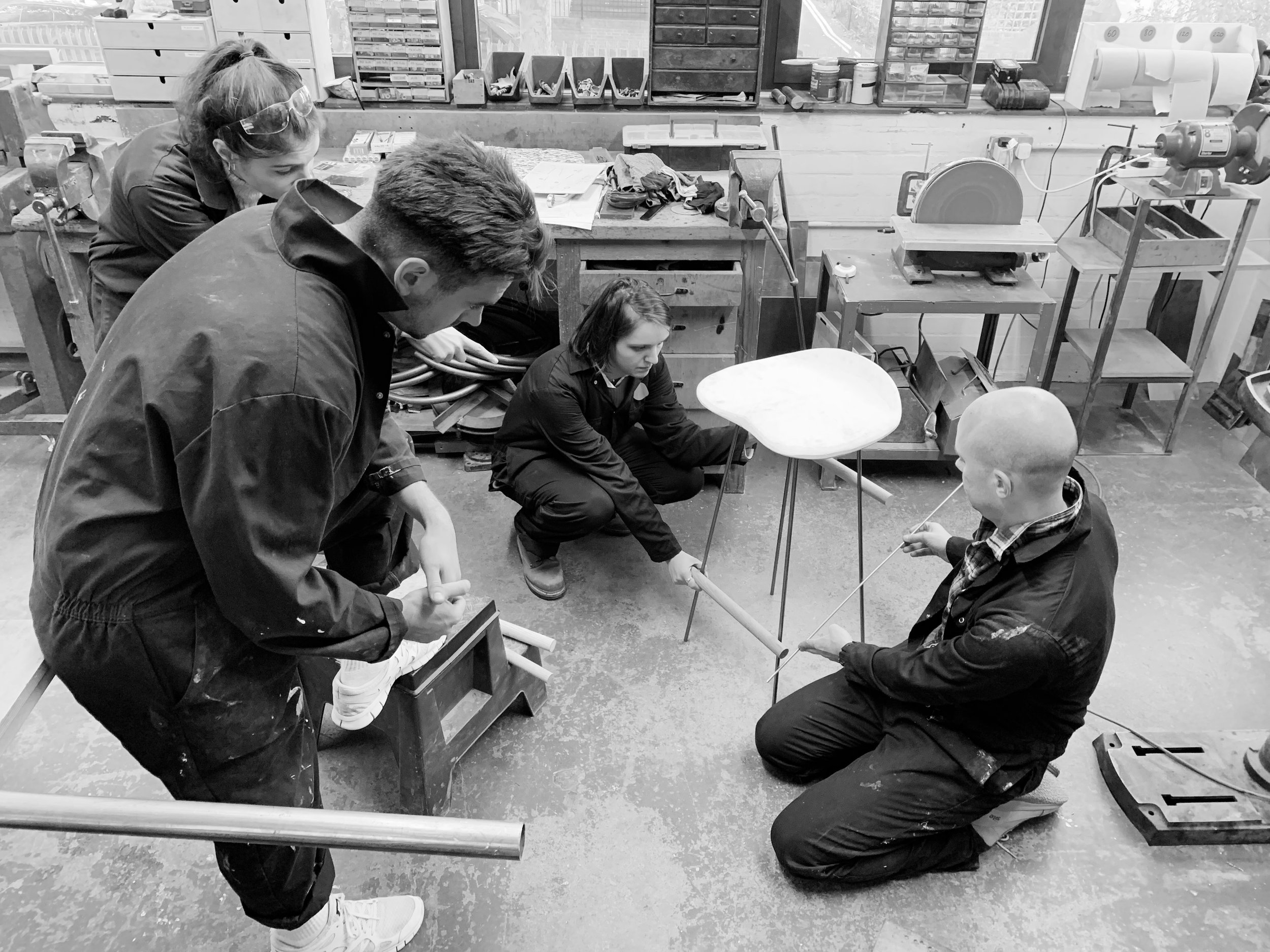 Four people in a workshop. Two seated, one kneeling, one standing, working on a project. Workshop equipment and tools are visible around.