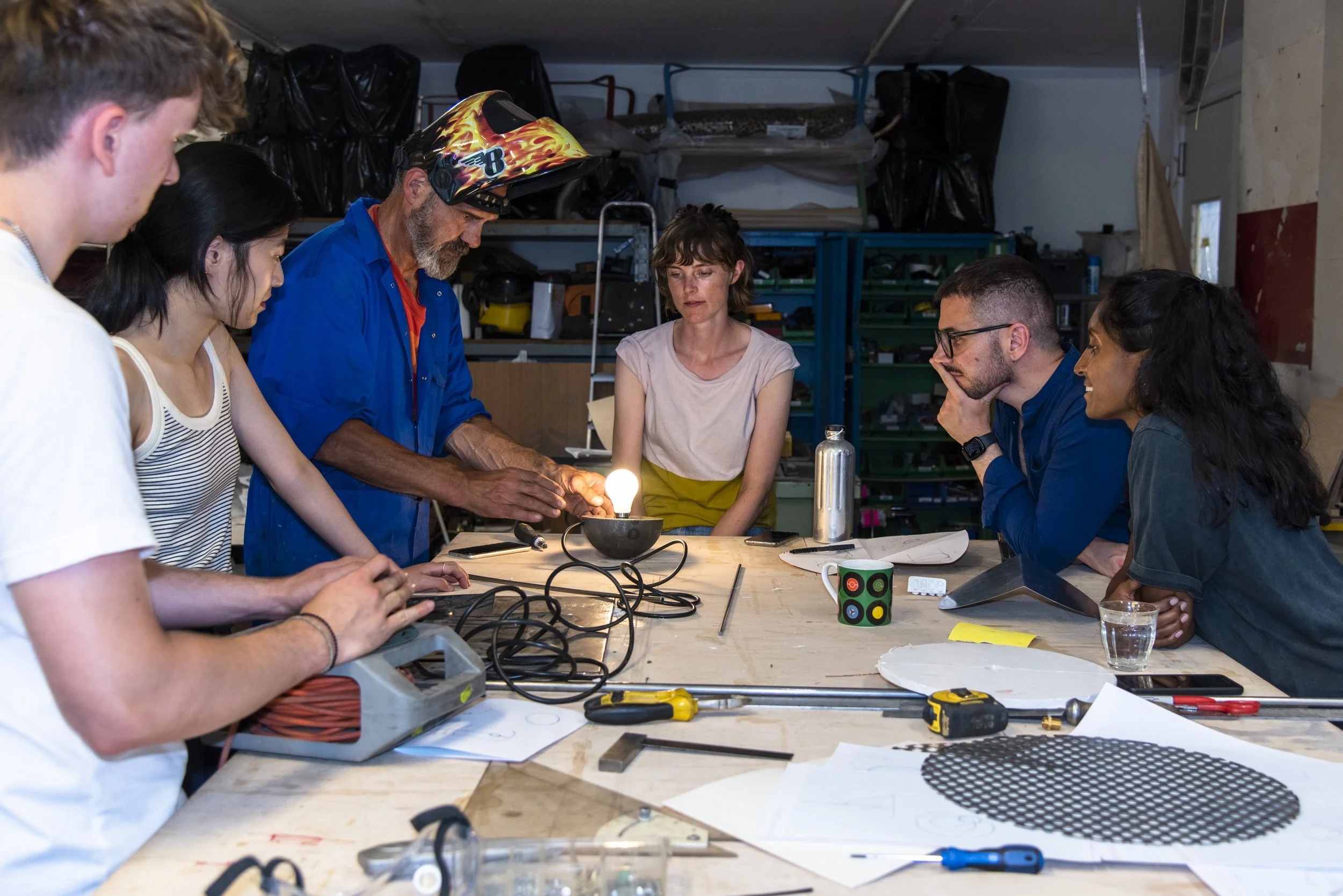 A group of people gathered around a workshop table, observing a man wearing a motorcycle helmet, working on a project with various tools and electronic components on the table.