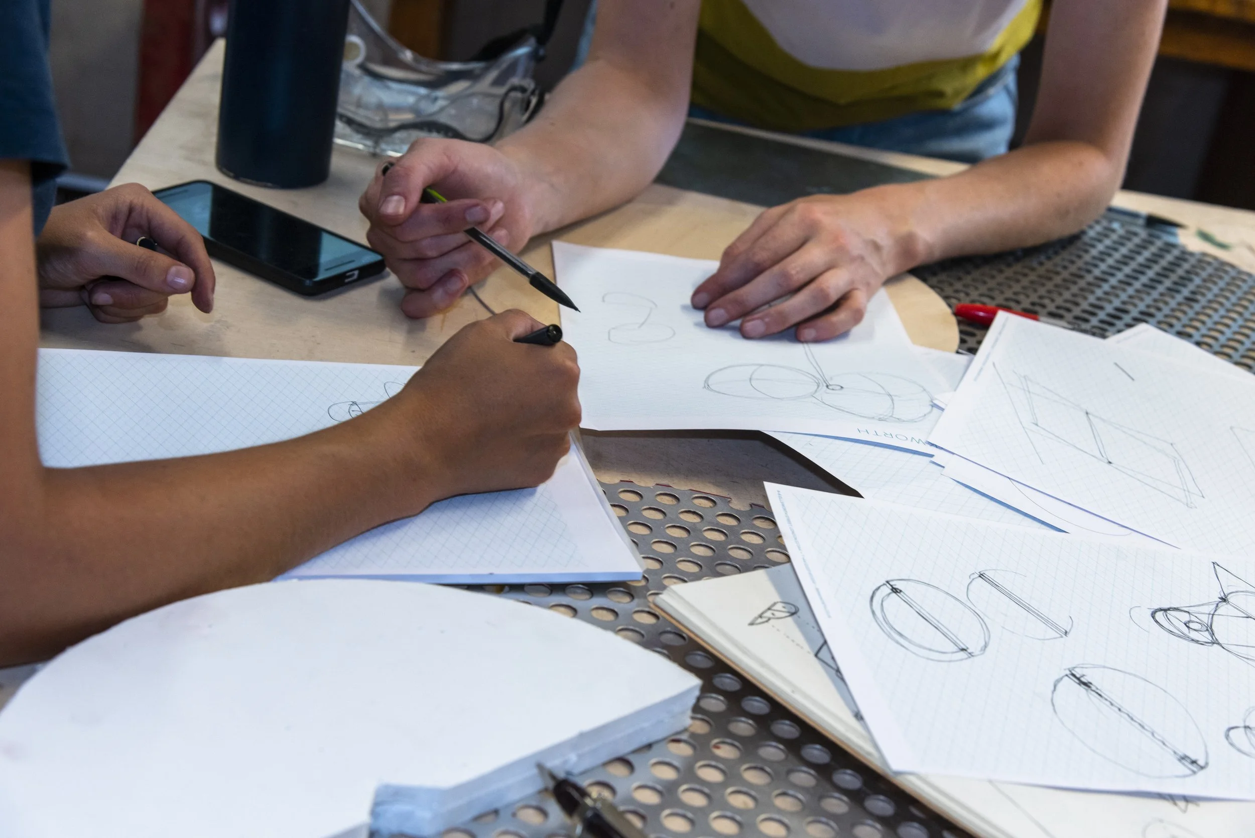 Two people working on drawing sketches at a table with papers, a smartphone, and writing utensils, including a black pen. One person is holding a pen, and the other is pointing or gesturing towards the papers.