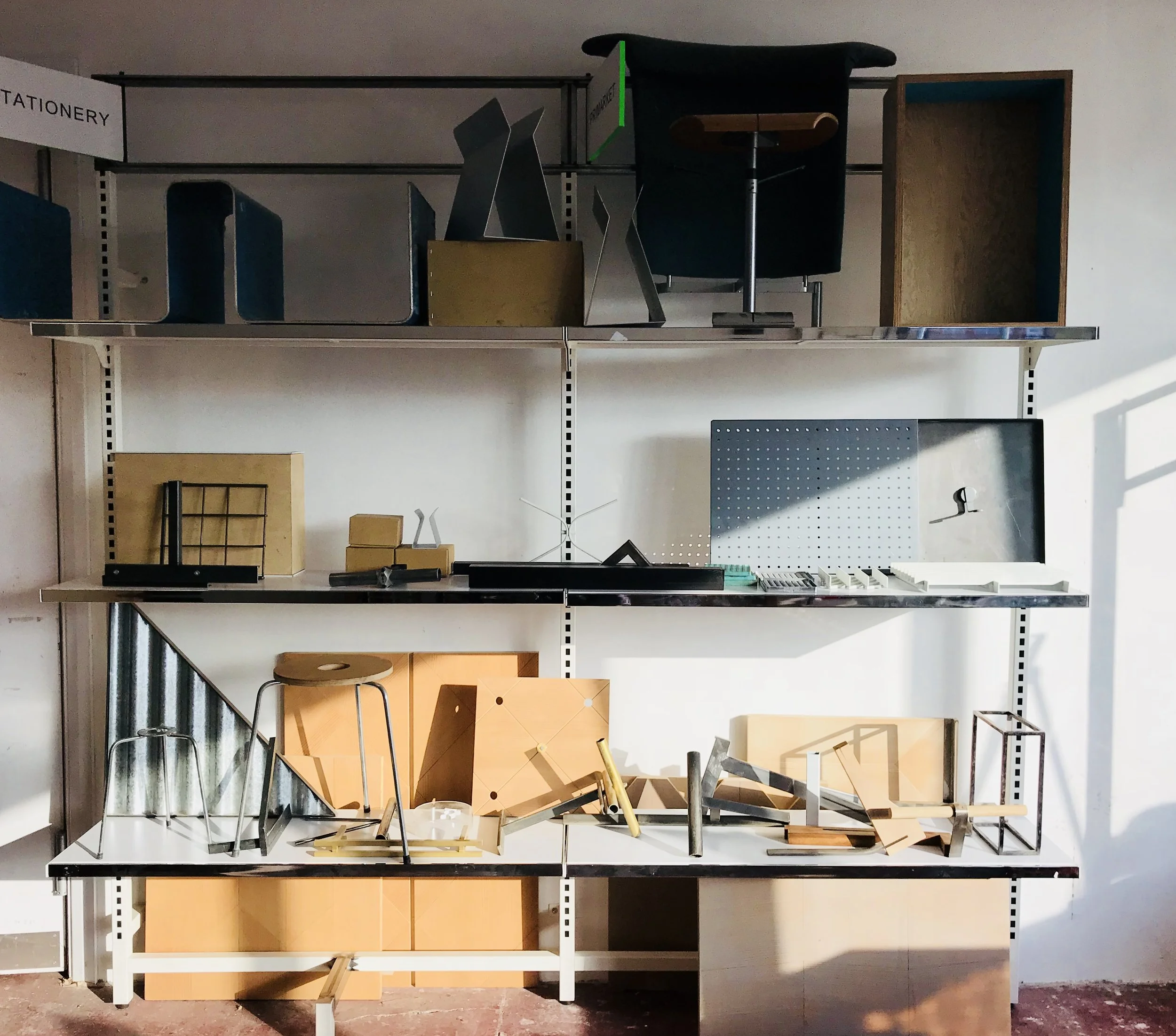 A display shelf with various office and home decor items, including chairs, boxes, and lighting fixtures, against a white wall with a wooden floor.