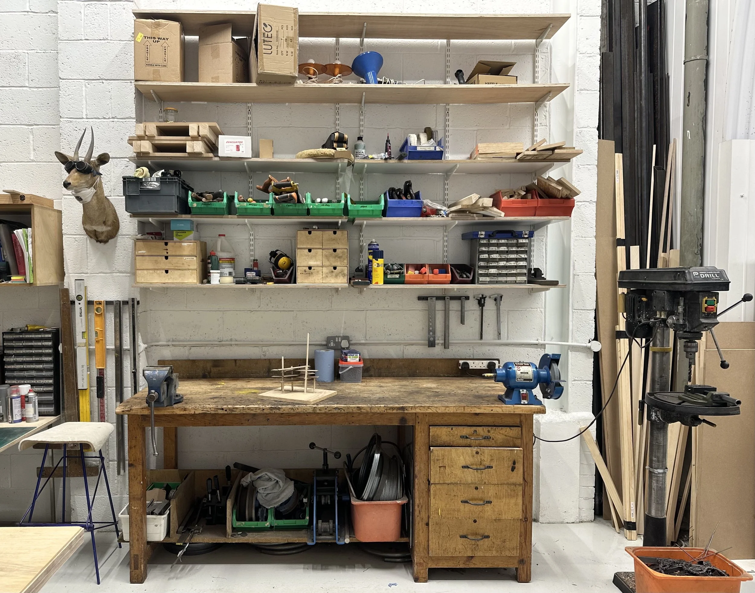 View of a woodworking workshop with a white brick wall, wooden workbench, and shelves holding various tools, supplies, and materials, including a mounted deer's head with sunglasses and a mask, and a drill press.