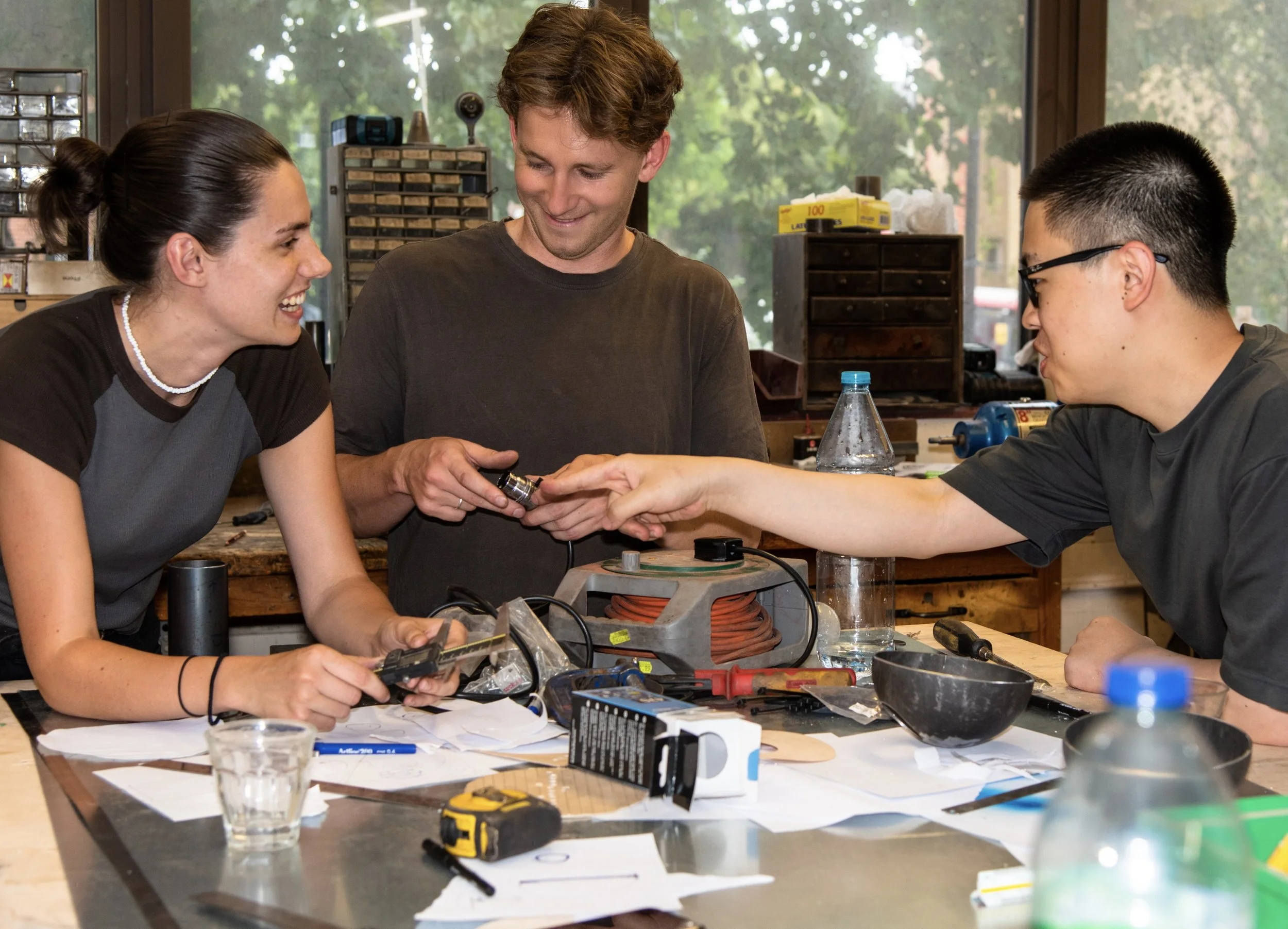 Three people working together in a workshop or lab, smiling and engaging with each other. They are surrounded by tools, electronic components, and papers on the table.