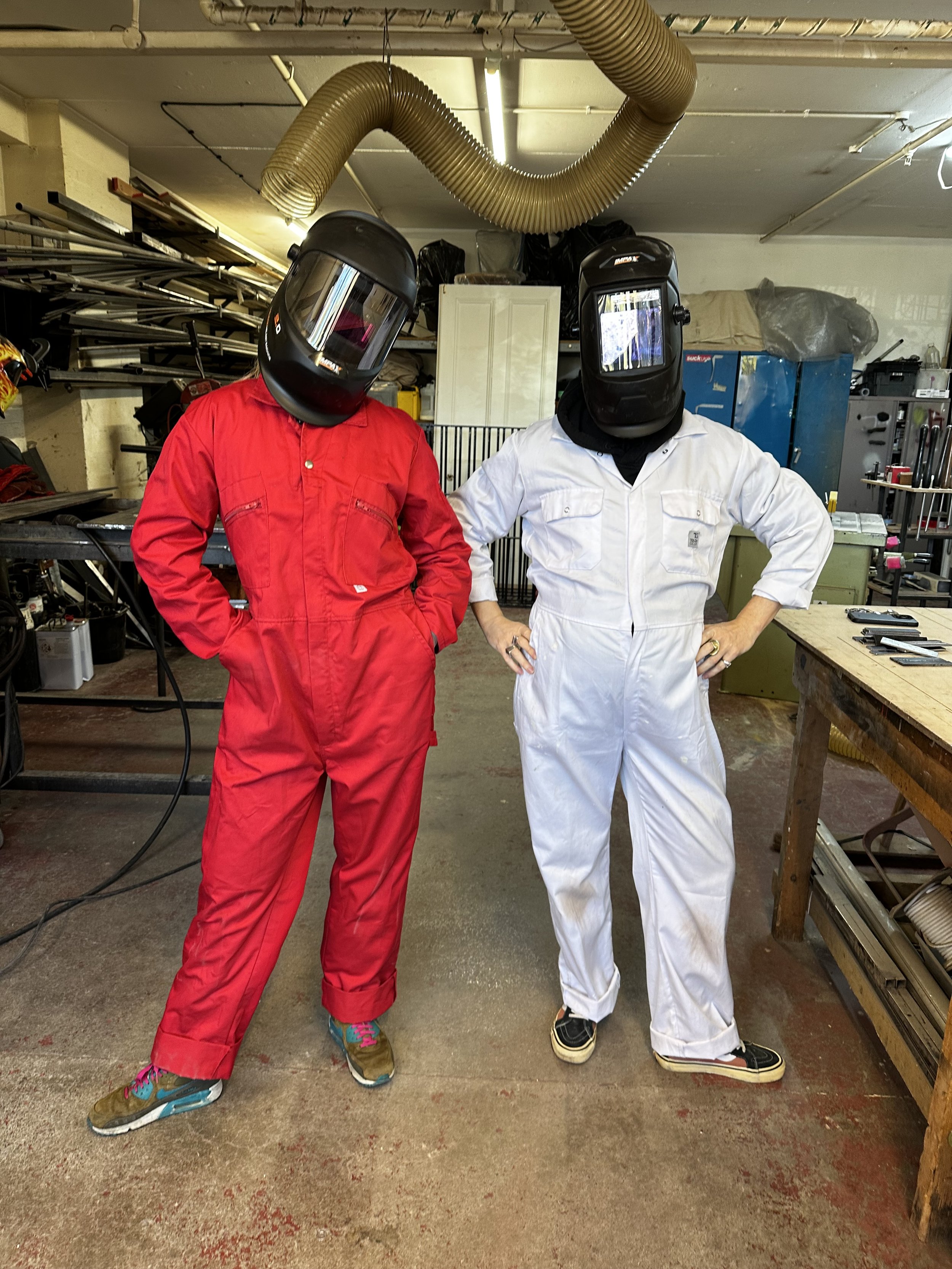 Two people are standing in a workshop or garage, wearing welding helmets and colorful jumpsuits (one in red, the other in white). They are posing with their hands on their hips. The workshop has tools, equipment, and storage shelves in the background.