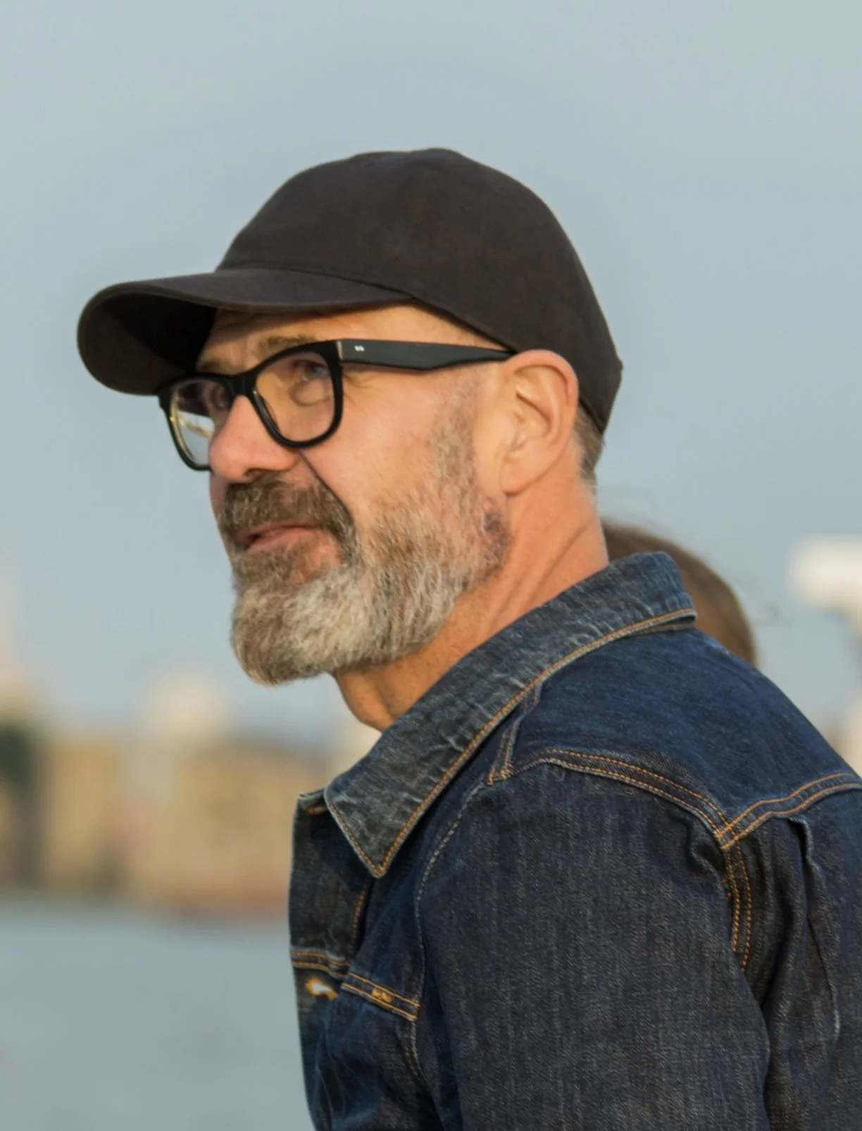 A middle-aged man with gray beard and glasses, wearing a black cap and denim jacket, looking to the side outdoors with a blurred background.