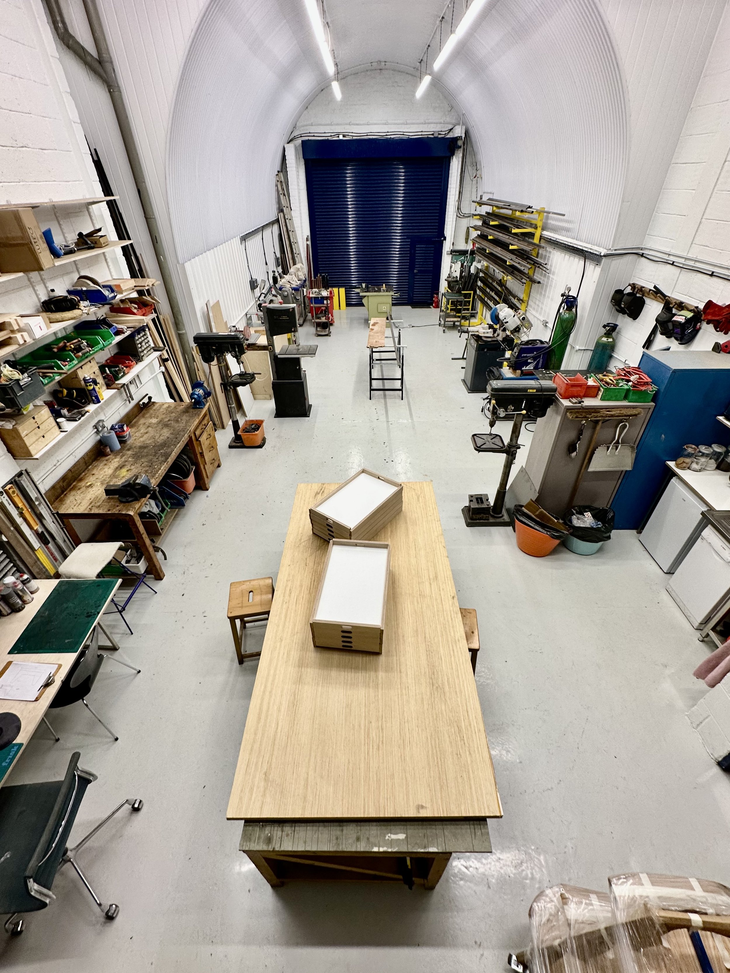 A workshop with a long wooden workbench in the center, various tools and equipment on shelves and tables along the walls, and a large blue roll-up door at the back.
