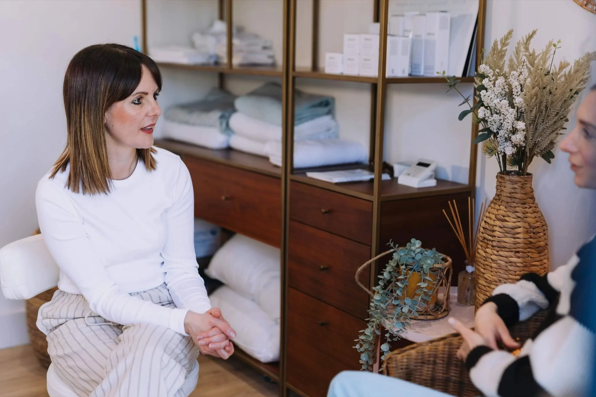 Two women engaged in conversation in a cozy room, with one sitting on a white chair and the other partially visible, surrounded by shelves with folded linens and decor items.