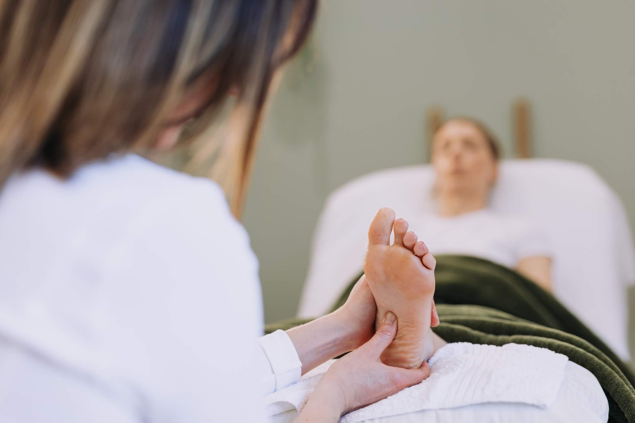 A person lying on a treatment bed receiving foot reflexology treatment, with a reflexologist holding the person's foot