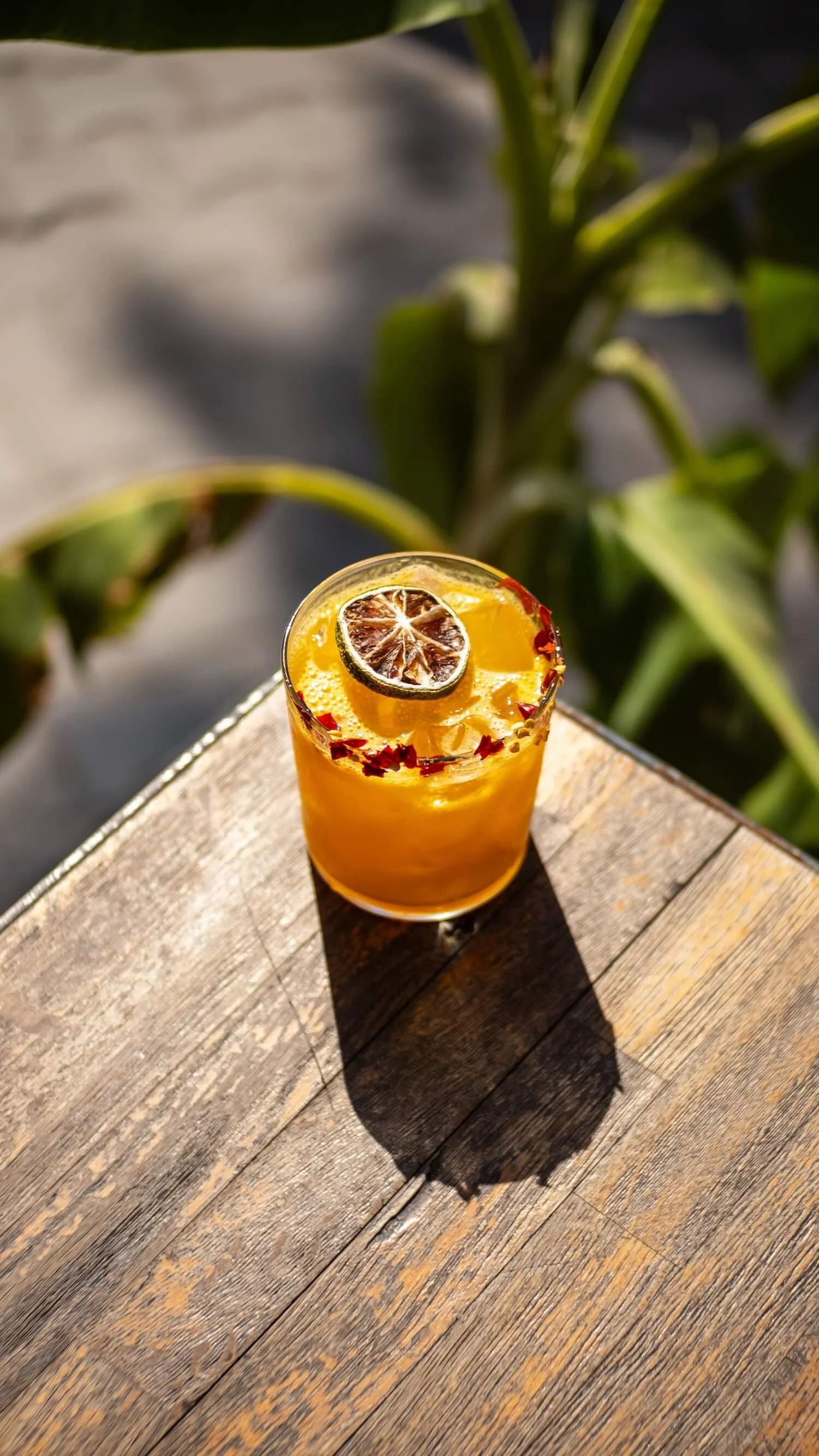 An orange margarita in a glass, garnished with a slice of dried lime and red chili flakes on the rim, on a rustic wooden table outdoors