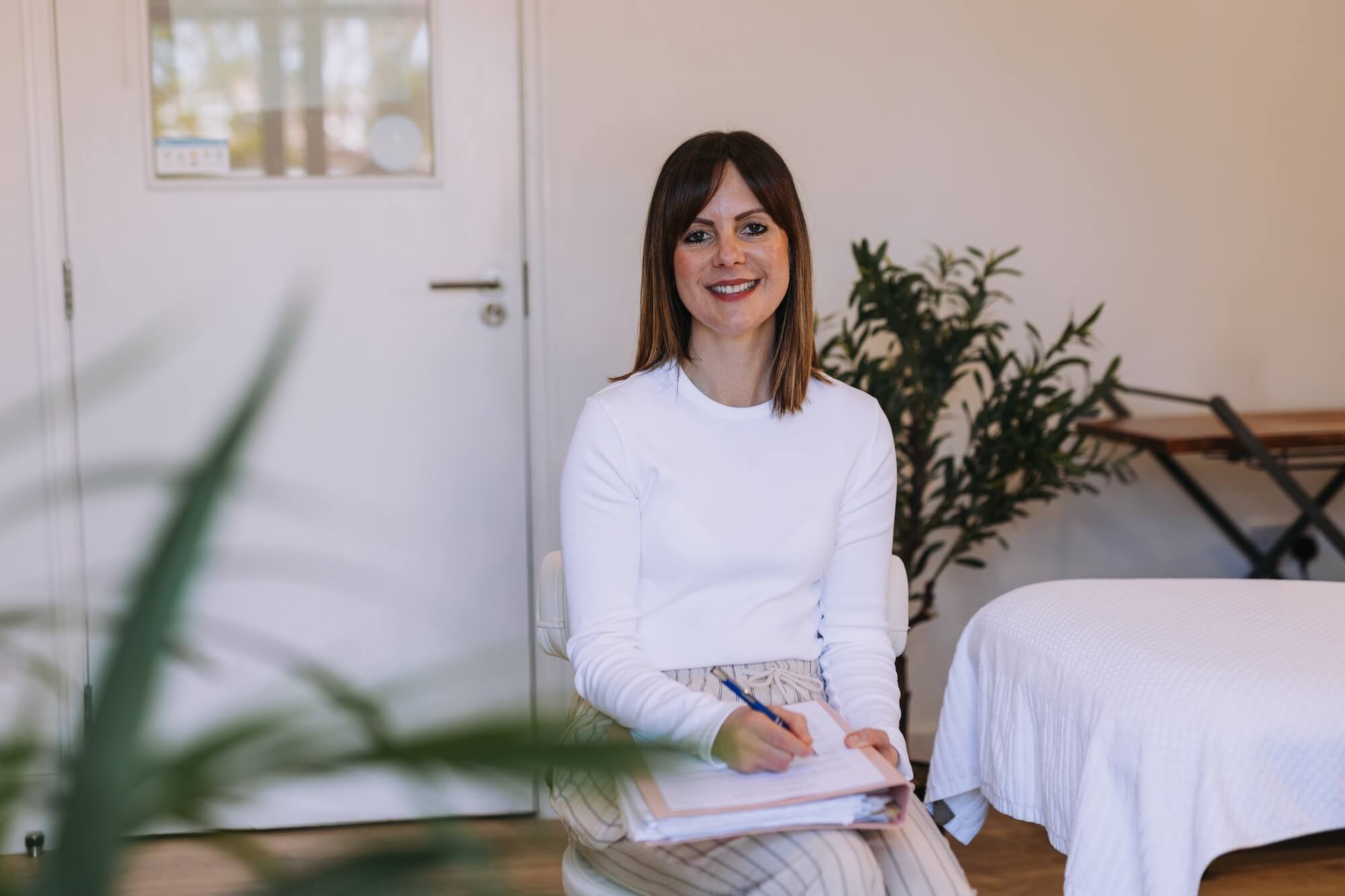 A woman with shoulder-length brown hair, wearing a white long-sleeve top, sitting on a chair in a bright room, holding a pen and notepad, with a large plant and a reflexology bed in the background