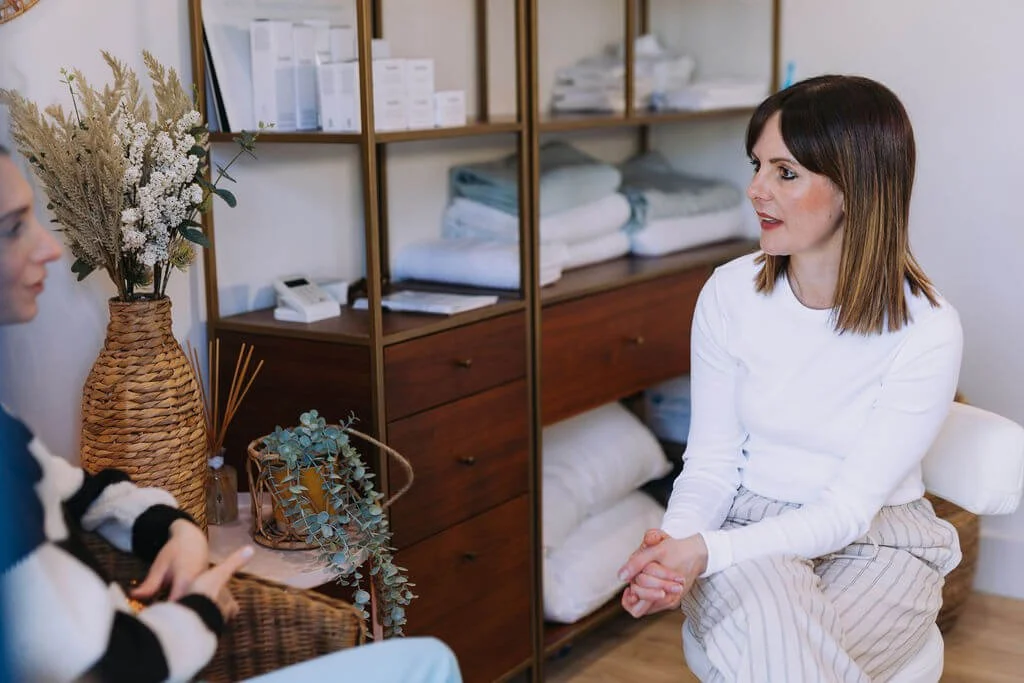 A woman in a white top with shoulder-length brown hair sitting and talking to another woman in a cosy, well-organized room with shelves of folded linens and a vase of dried flowers