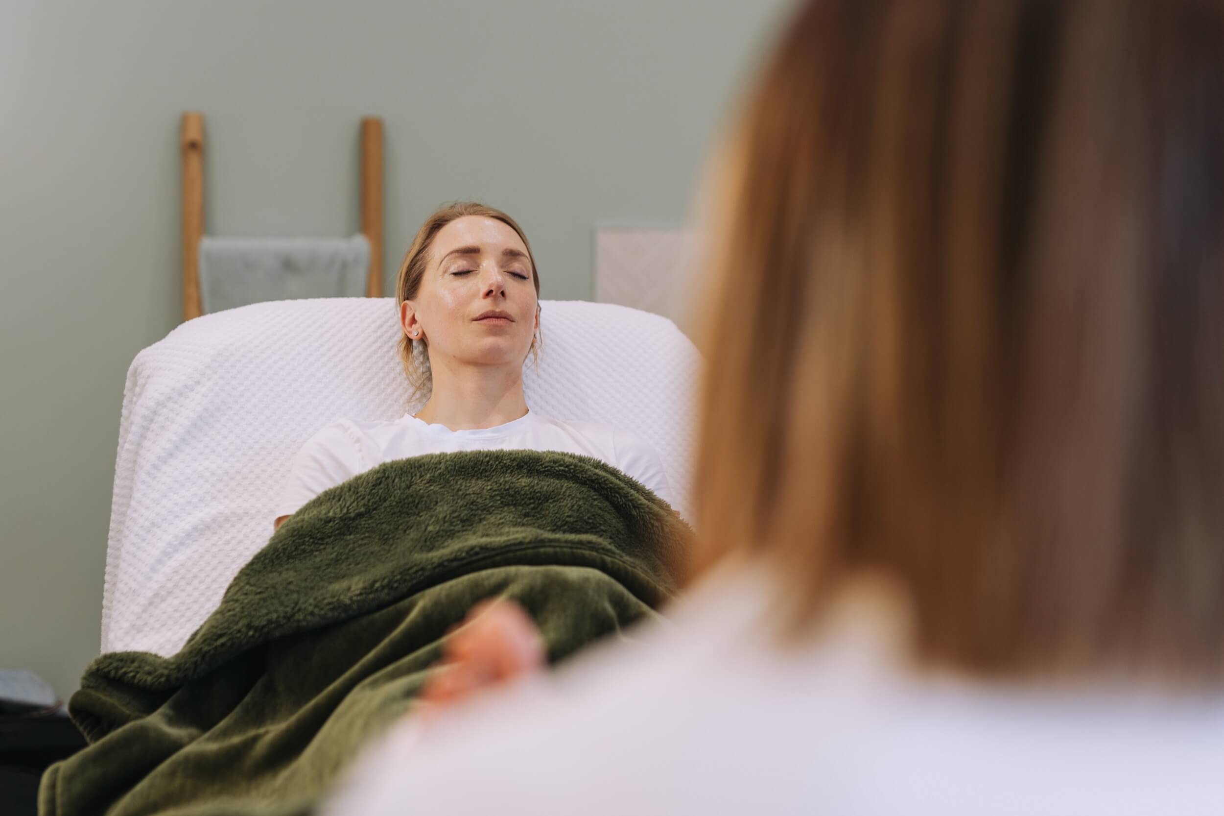 A woman with closed eyes lying on a bed, covered with a green blanket, receiving foot reflexology treatment