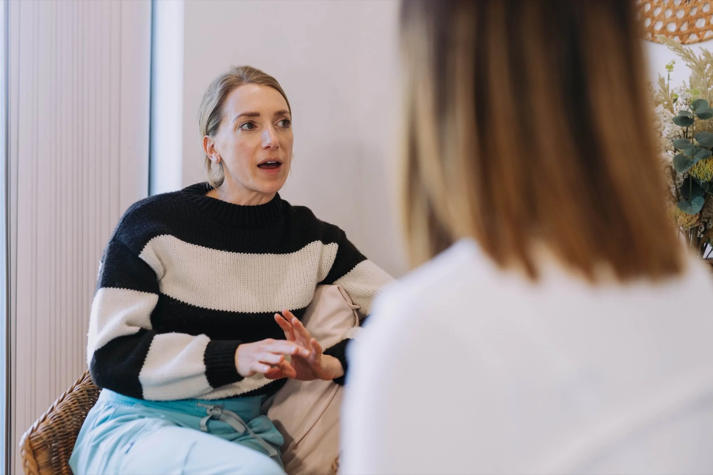 A woman with blonde hair, wearing a black and white striped sweater, is sitting and talking to another person whose back is visible, showing brown hair. The setting appears to be indoors with a light background and decorative plants on a table.