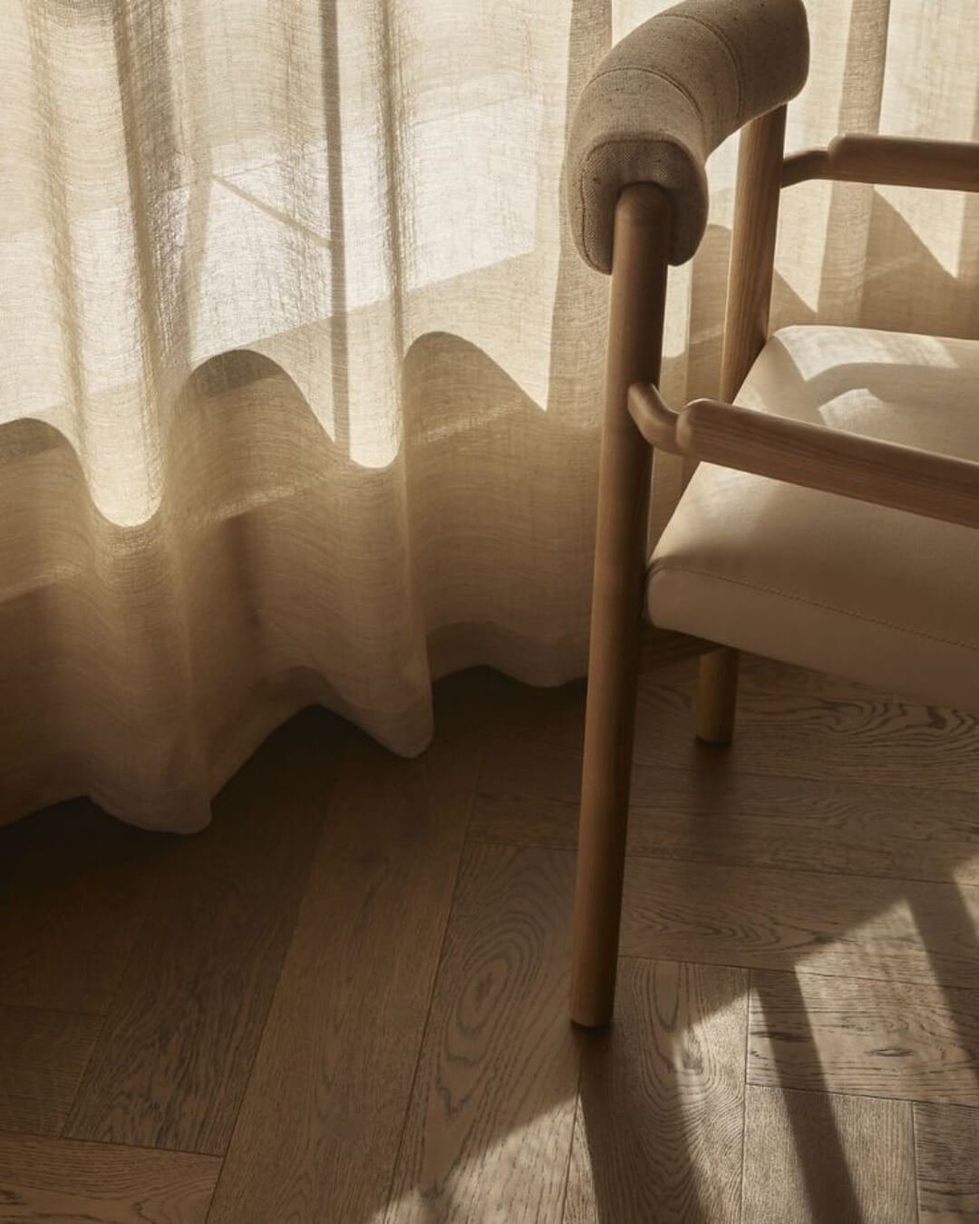 Sunlight filters through sheer curtains, casting shadow patterns on a wooden floor next to a light-coloured upholstered chair with wooden arms