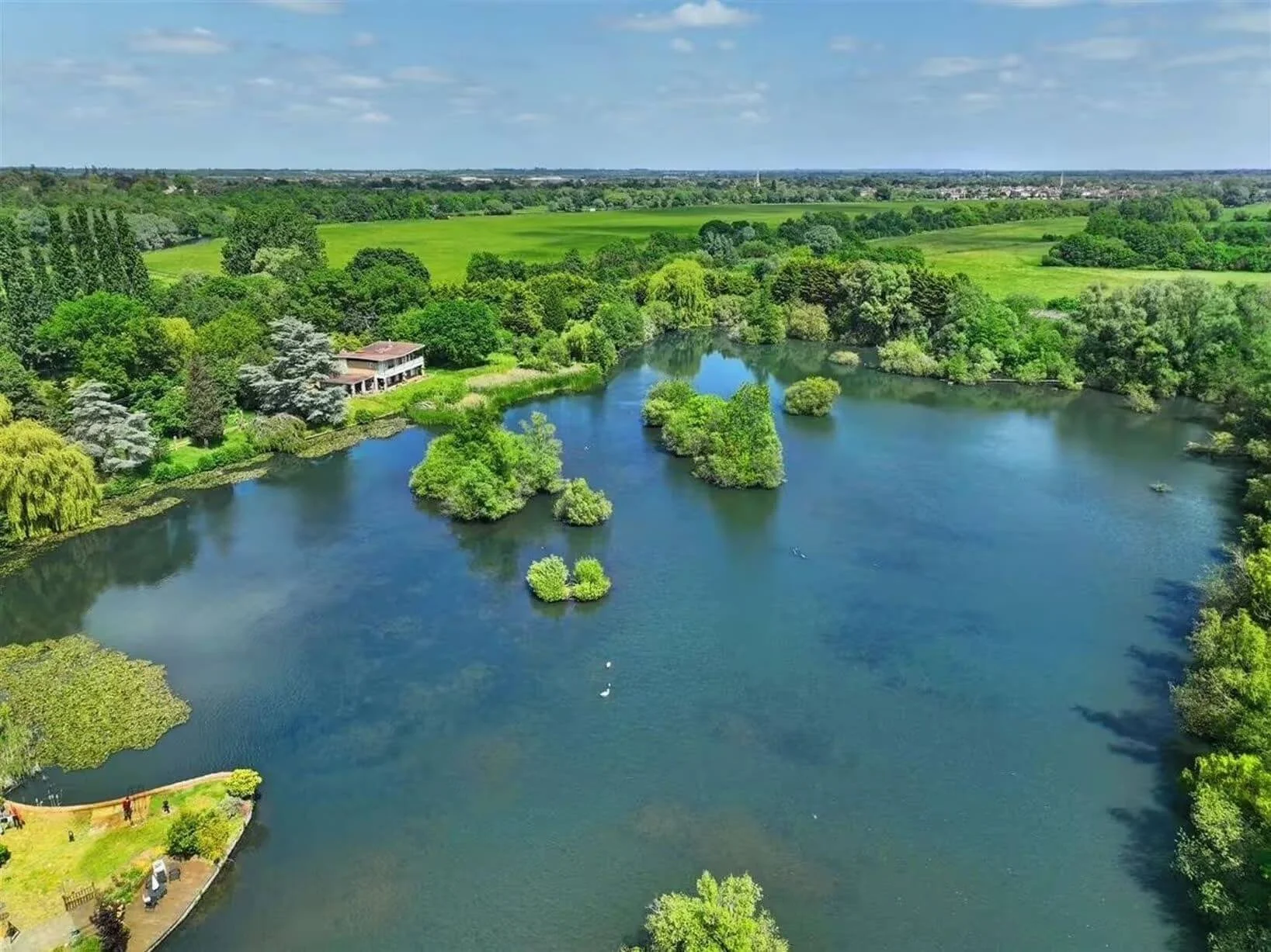 Aerial view of a large pond surrounded by lush green trees and fields, with a house on the left side and small boats on the water under a partly cloudy sky.