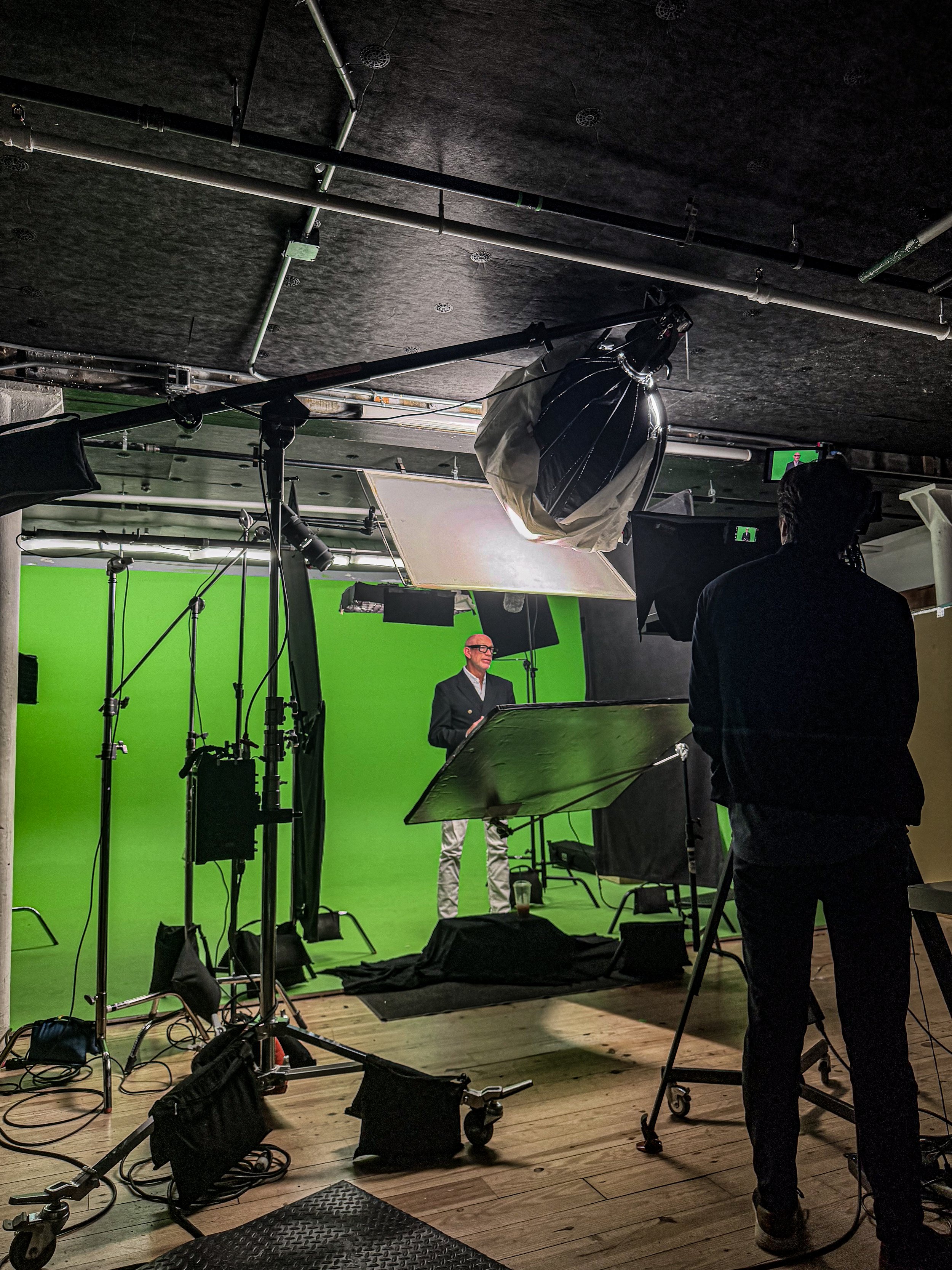 A behind-the-scenes view of a video shoot in a studio with a green screen, lighting equipment, and a person in a suit being filmed.