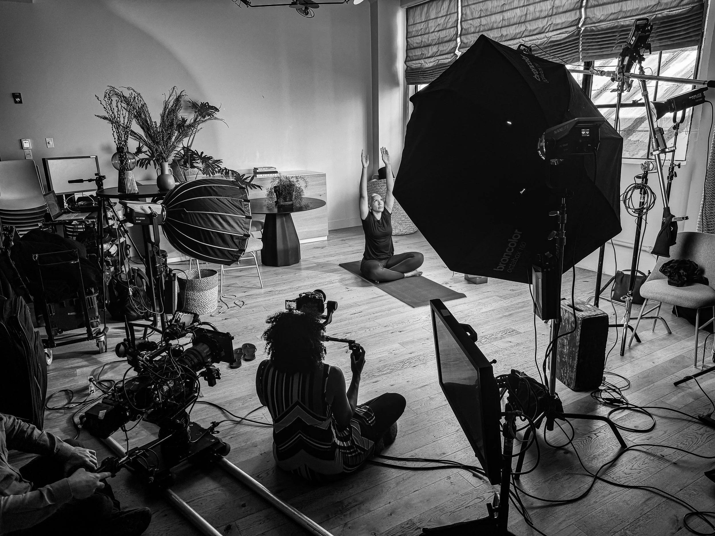 A woman practicing yoga on a mat in a room with photography equipment and lighting, with large window blinds and plants in the background.