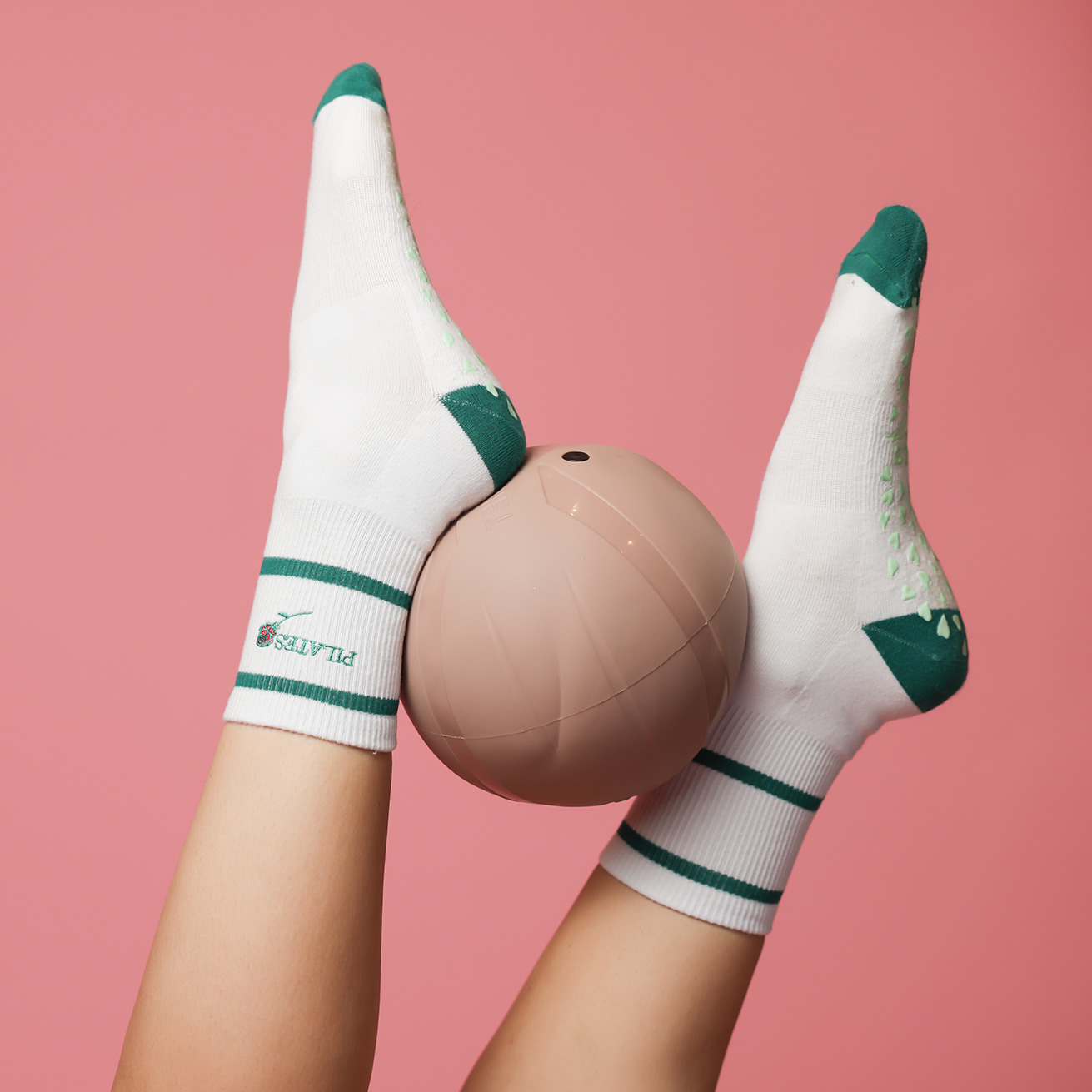 A person wearing white socks with green tips, holding a pink volleyball against a pink background.