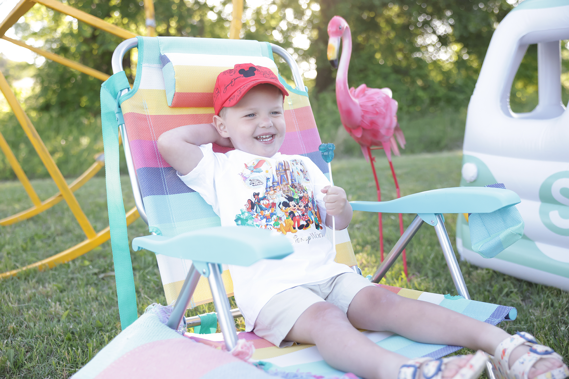 A young boy sitting in a colorful outdoor stroller with a rainbow-striped canopy, smiling and wearing a red cap and a Disney-themed t-shirt, with a pink flamingo lawn ornament and a white and blue toy vehicle in the background on grass with trees.