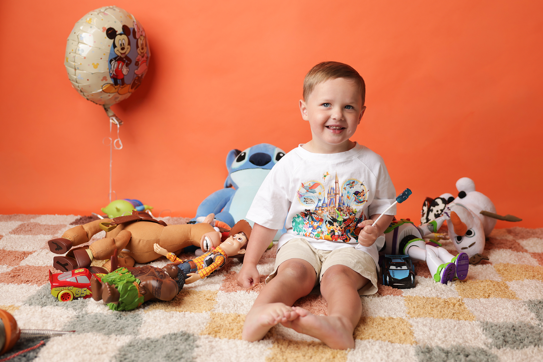 A young boy with blonde hair and a Disney-themed t-shirt sitting on a colorful carpet surrounded by toys including plush characters, cars, and balloons against an orange background.