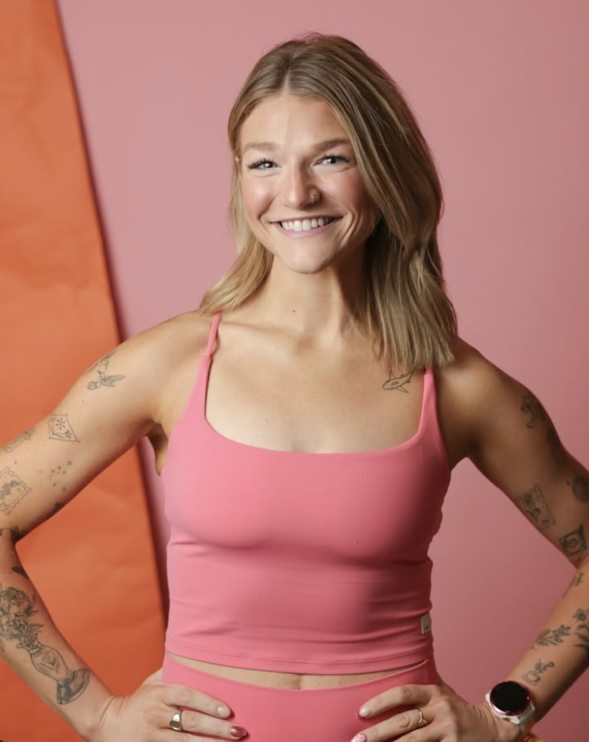 Smiling tattooed woman in a pink tank top and matching bottoms posing against a pink and orange background