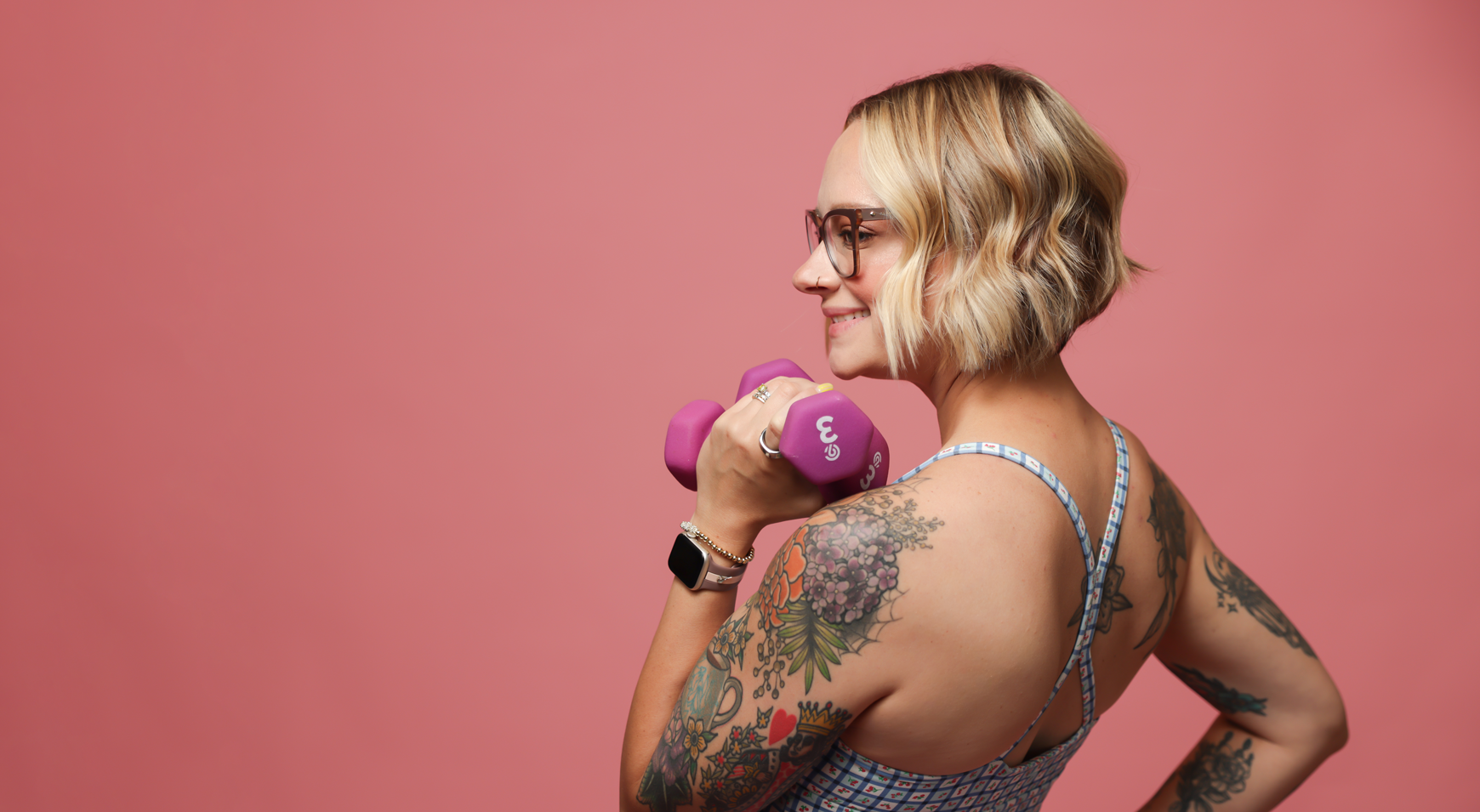 A woman with blonde wavy hair, glasses, and tattoos on her arm, holding a pink dumbbell against a solid pink background.