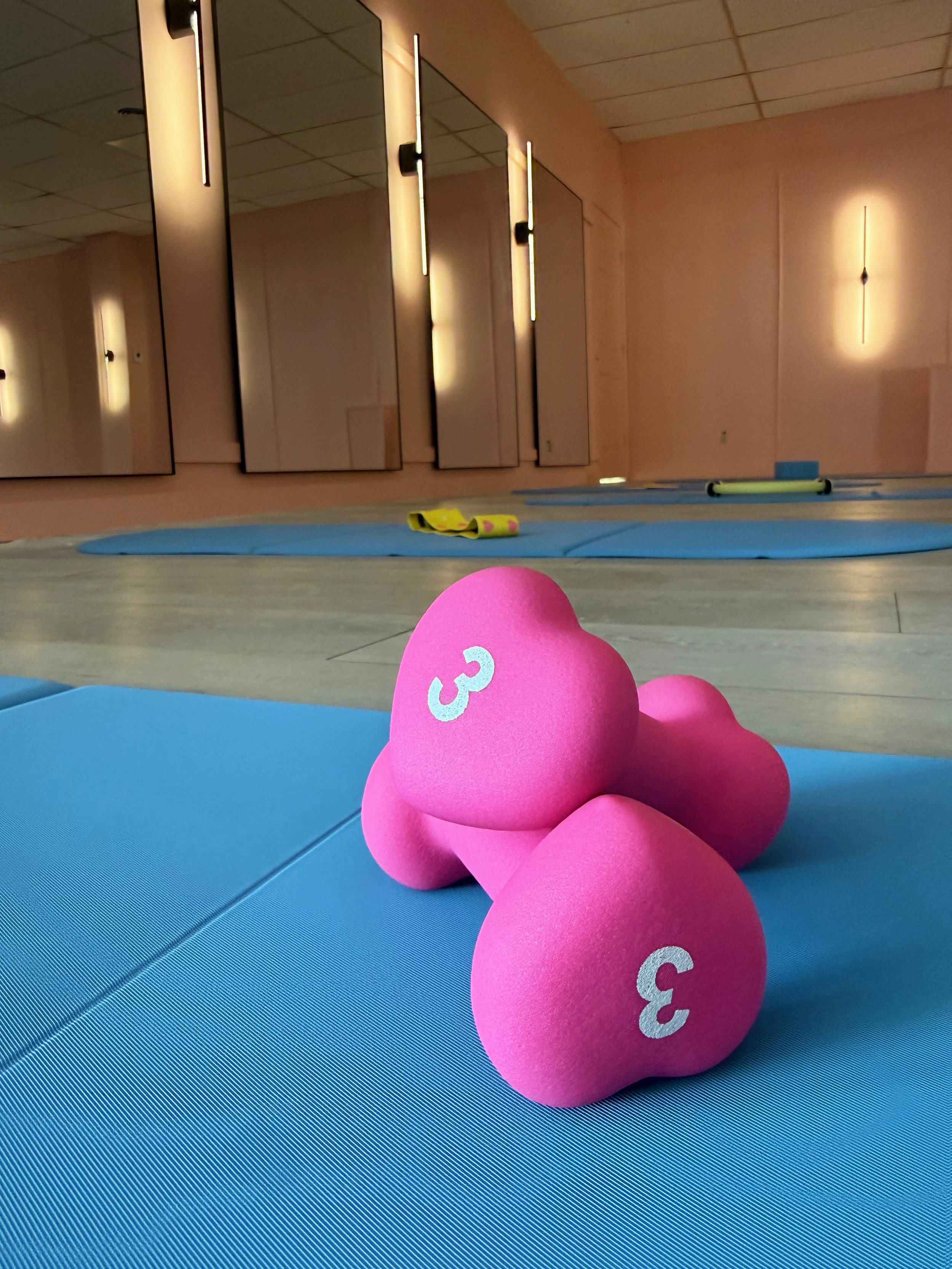 Pink hand weights with the number 3 on a blue exercise mat, in a fitness room with mirrors and wall lights.