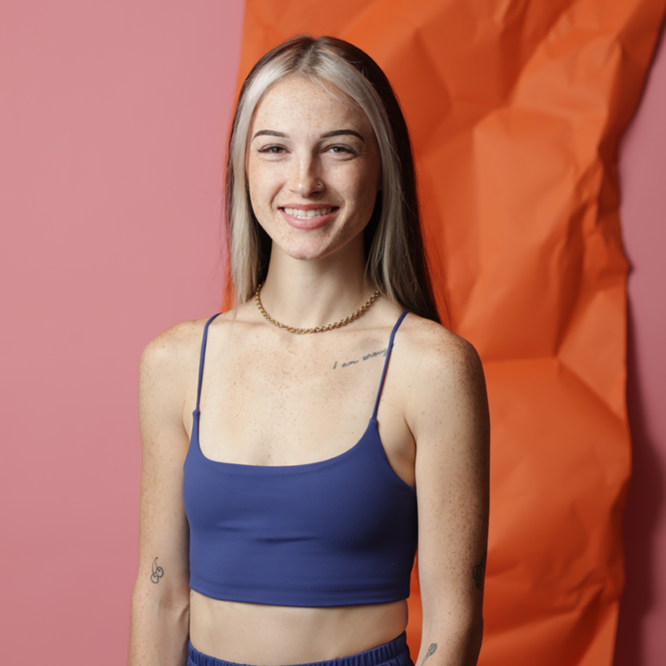 Young woman with blond hair, freckles, and tattoos, smiling and wearing a blue crop top and a gold chain necklace, standing against a pink and orange paper backdrop.
