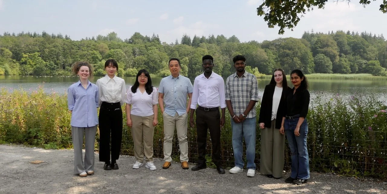 Group of eight diverse young adults standing outdoors by a lake with trees in the background.