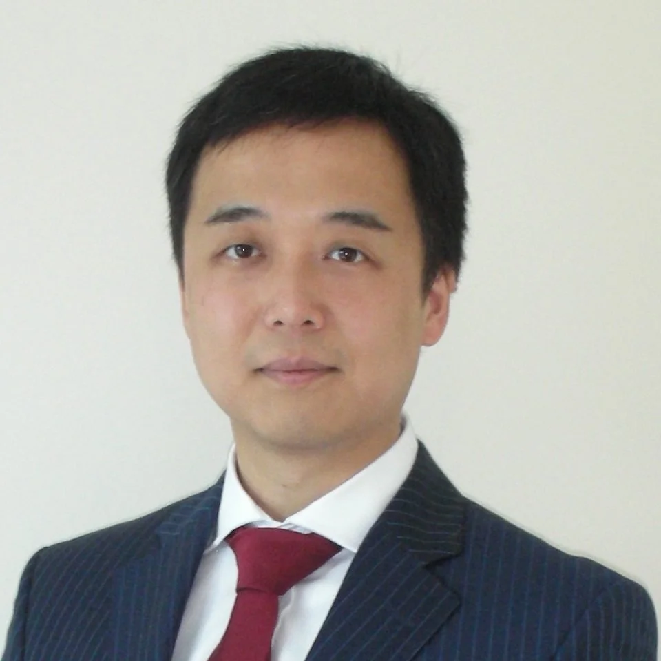 A man in a business suit with a white shirt and a red tie, standing against a light-colored background.