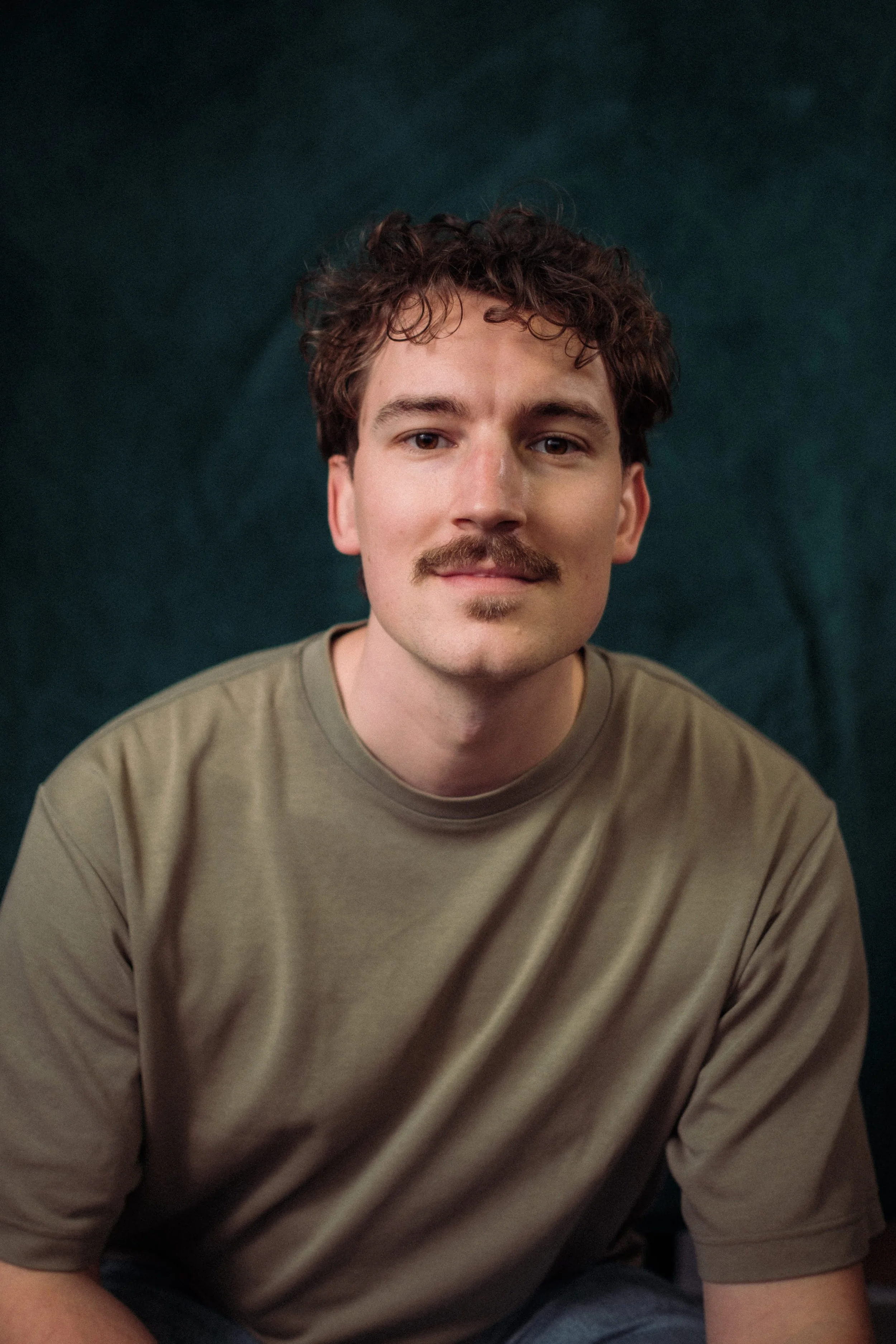 Portret van een jonge man met een lichte huid, kastanjebruine krullende haar en een snor, zittend met een donkere achtergrond.