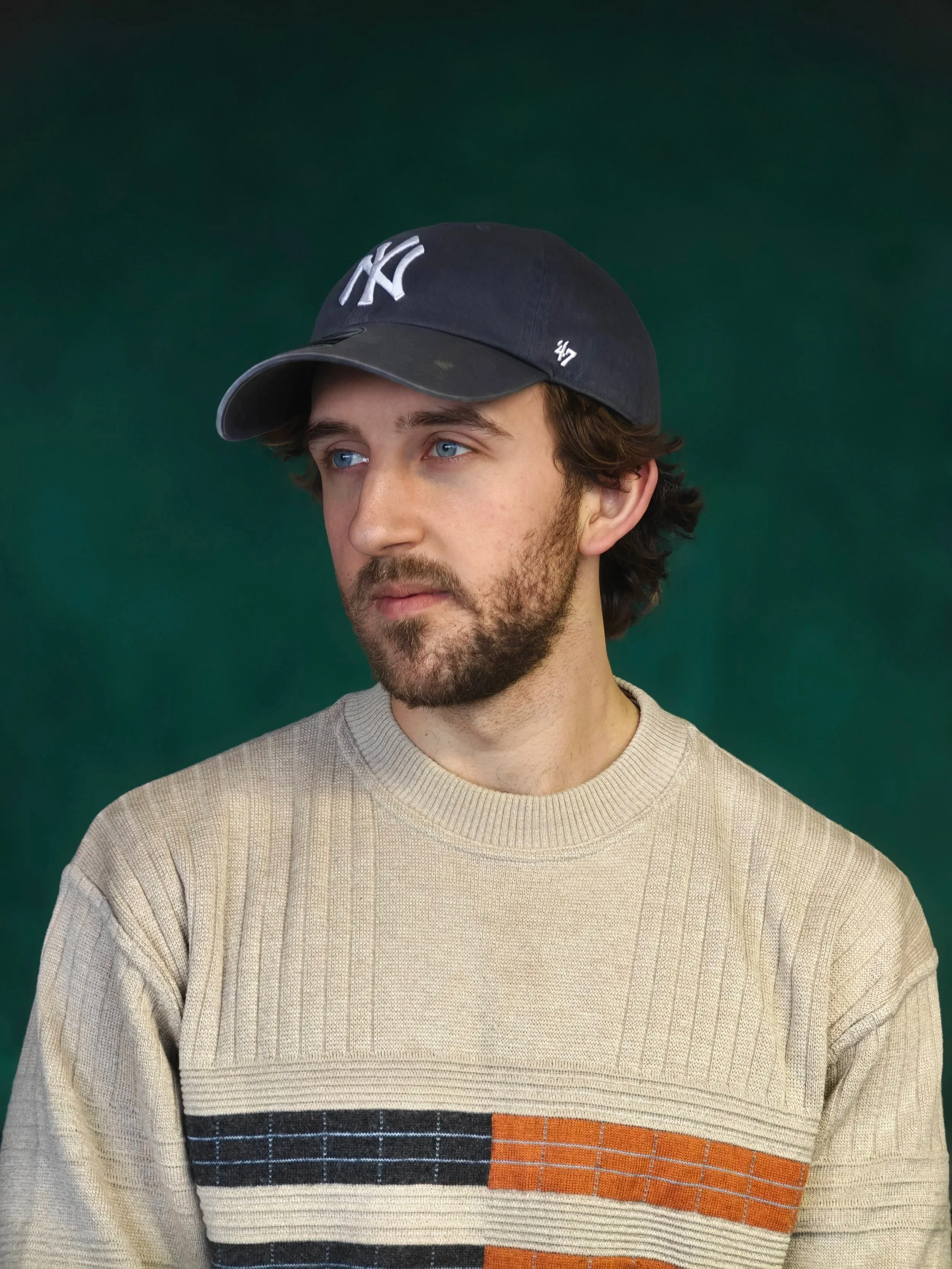 Een jonge man met blauwe ogen, een baard, en donzig, golvend haar, draagt een zwarte baseballpet met het witte logo van de New York Yankees en een beige trui met een ontwerp van zwart, oranje en grijs geruite patronen.