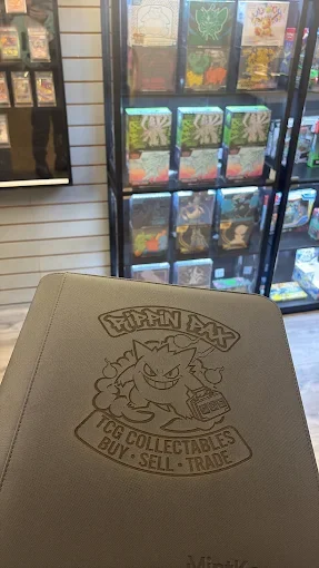 A Pokémon-themed notebook with a Greninja character holding a trading card on the cover, placed on a glass display case with various Pokémon cards in the background.