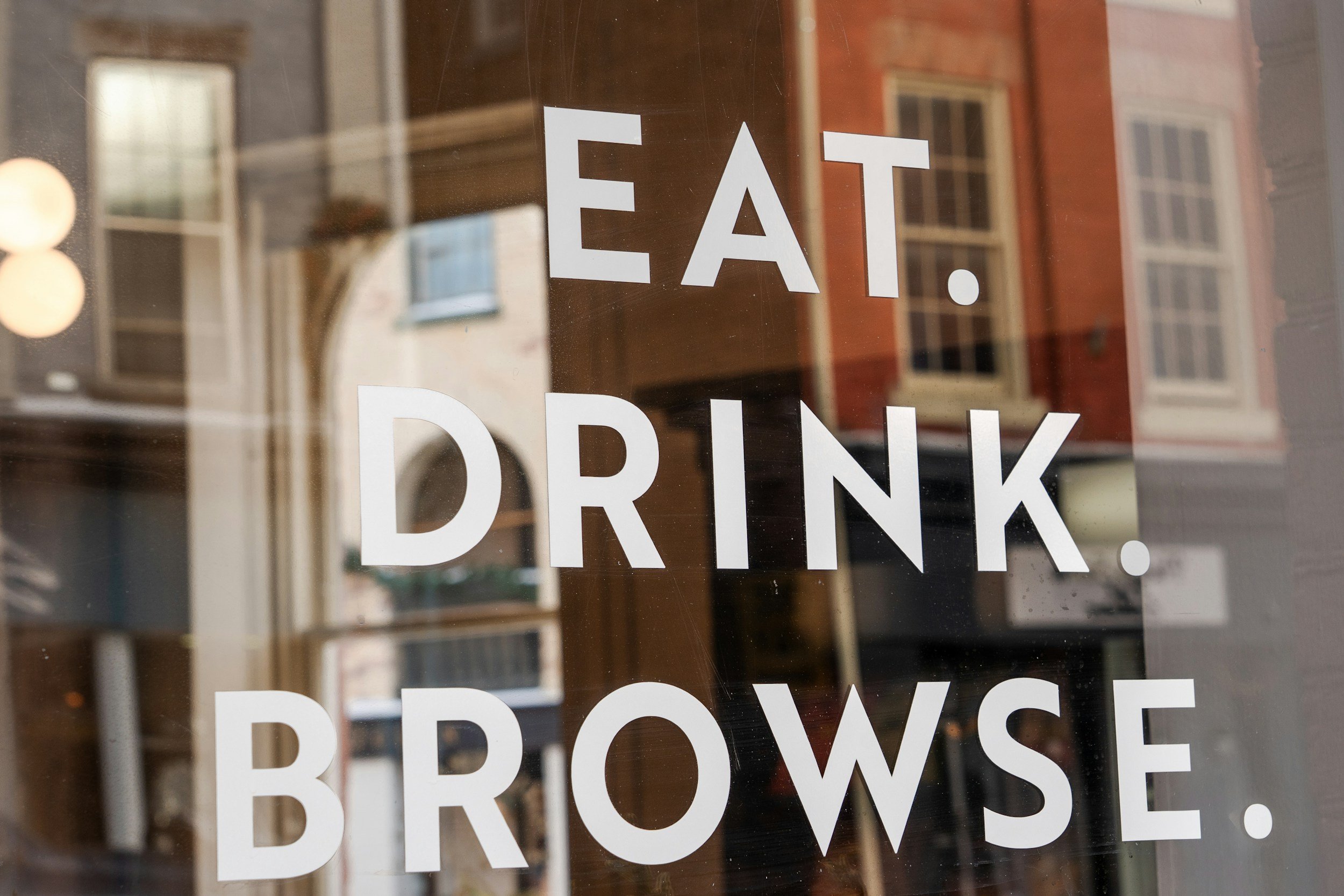 Close-up of a window with white text that reads 'EAT. DRINK. BROWSE.' Reflections of buildings and windows are visible on the glass.