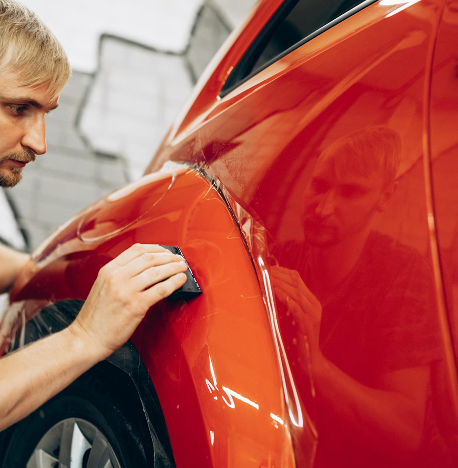 A man in a workshop repairing or inspecting the front quarter panel of a shiny orange car, with a scratch or minor damage visible.