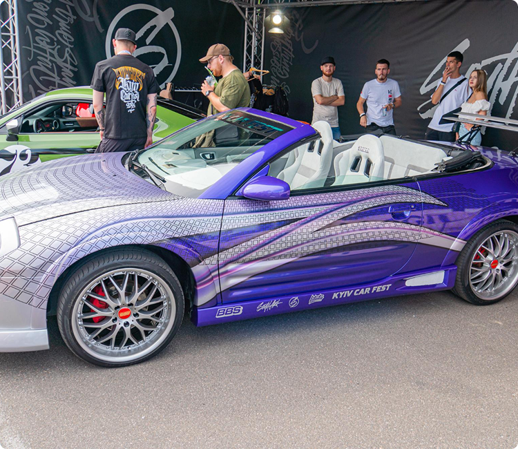A purple and silver convertible sports car on display at a car event with people gathered around.