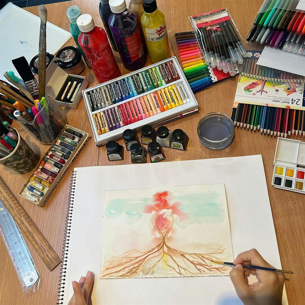 Overhead photo of hands painting a watercolor tree with spreading roots and a red, feathery canopy in a sketchbook, surrounded by oil pastels, paint bottles, colored pencils, ink jars, and brushes on a wooden table.