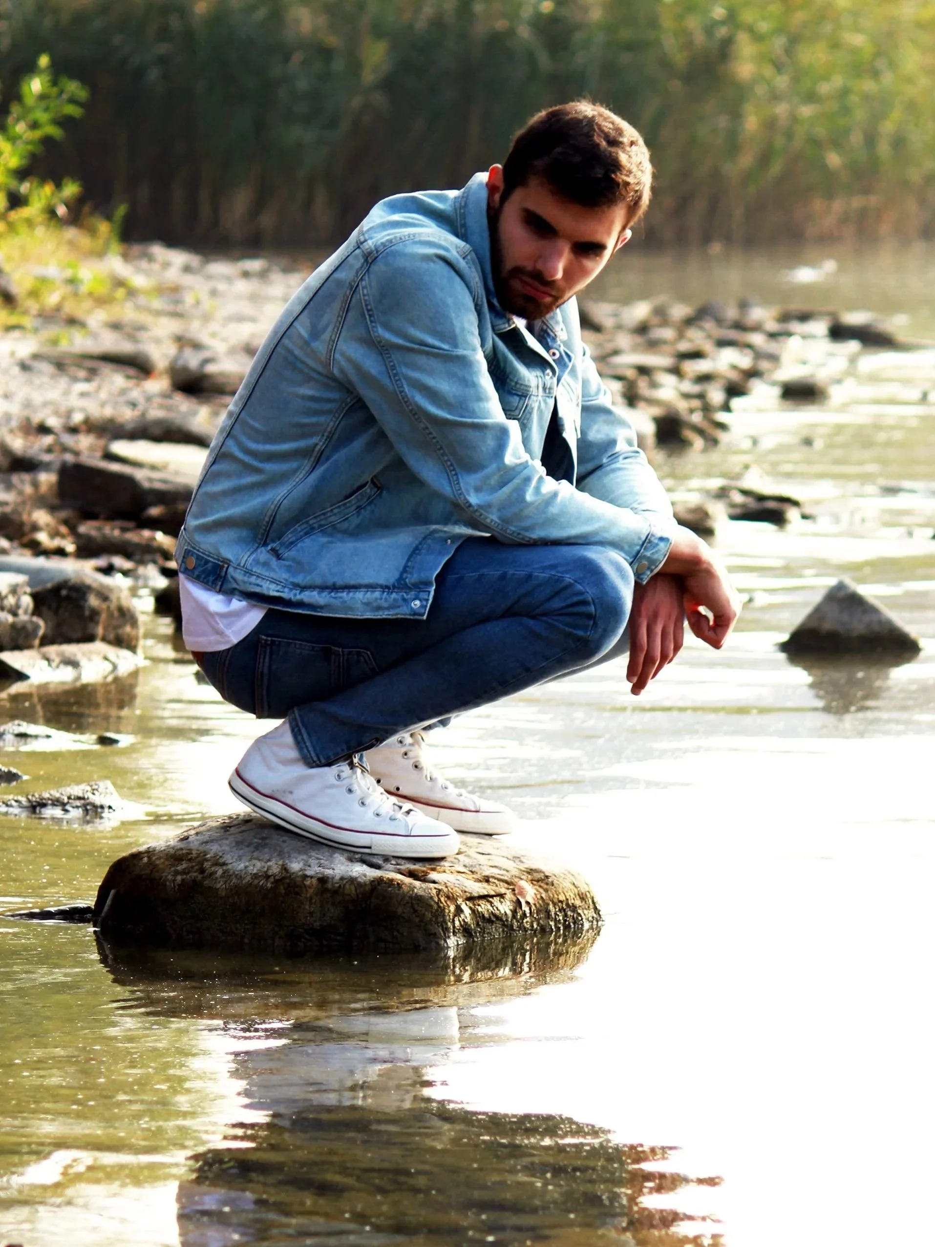 A man crouching reflectively on a rock near water, embodying quiet self-reflection and emotional grounding.