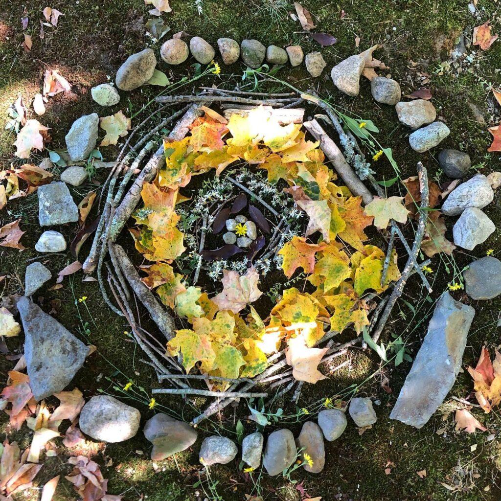 Overhead photo of a nature mandala on mossy ground: concentric circles of smooth stones, twigs, and yellow and orange autumn maple leaves, with small stones and a tiny yellow flower at the center.