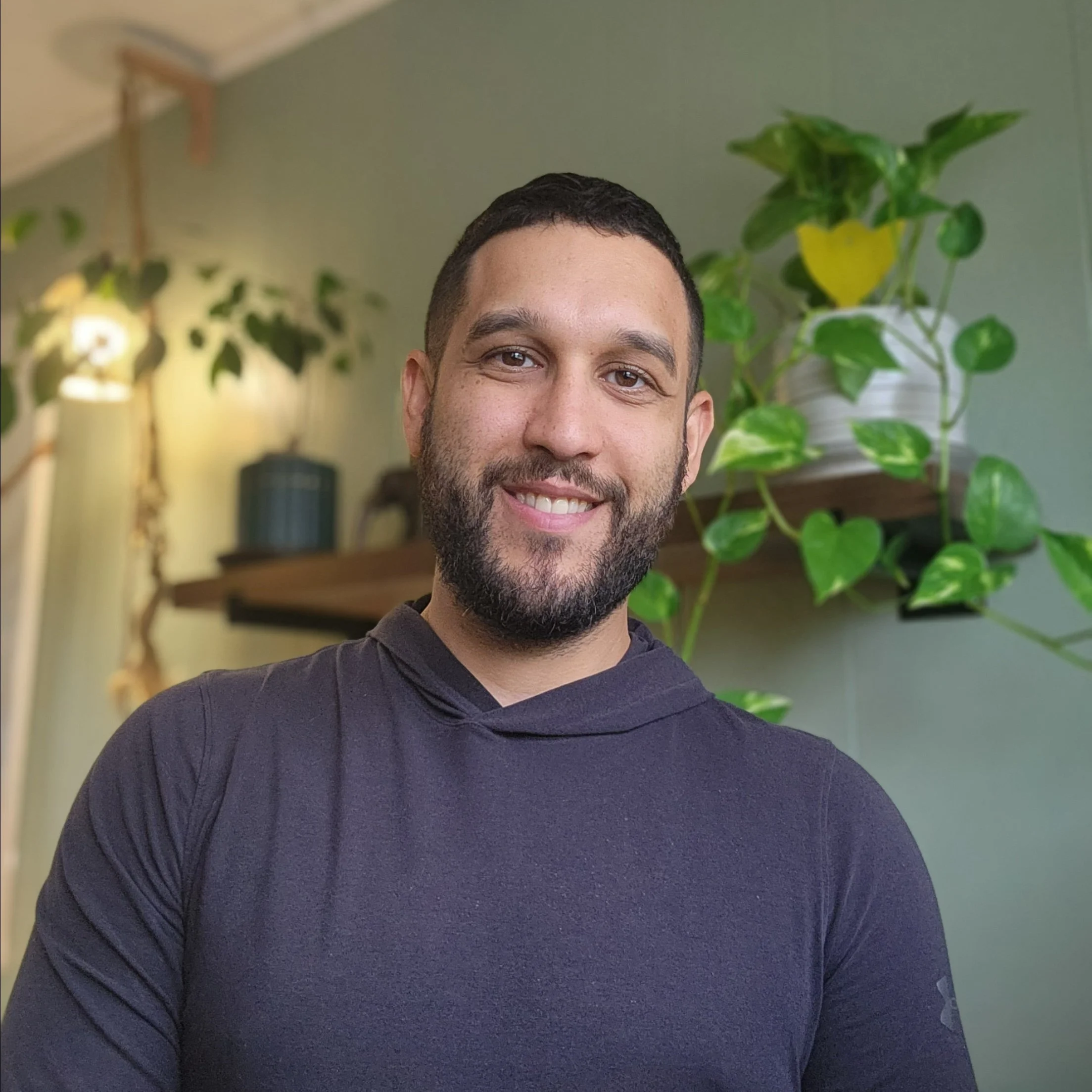 Close-up of a smiling man with short dark hair and a beard, wearing a dark blue sweater, standing in front of green houseplants and a green wall