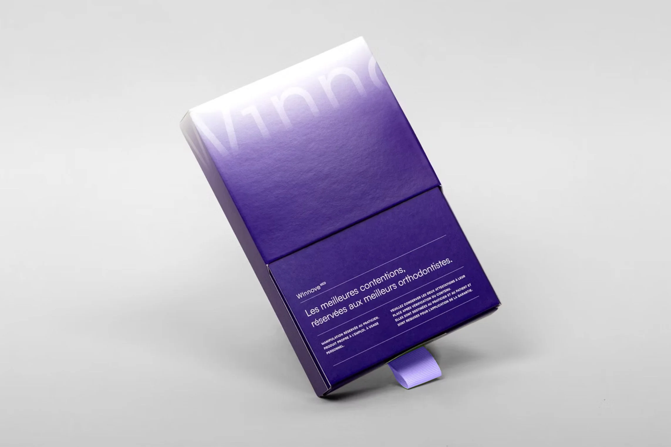 Close-up of a purple box with French text, featuring a ribbon at the bottom, positioned on a plain light grey surface.
