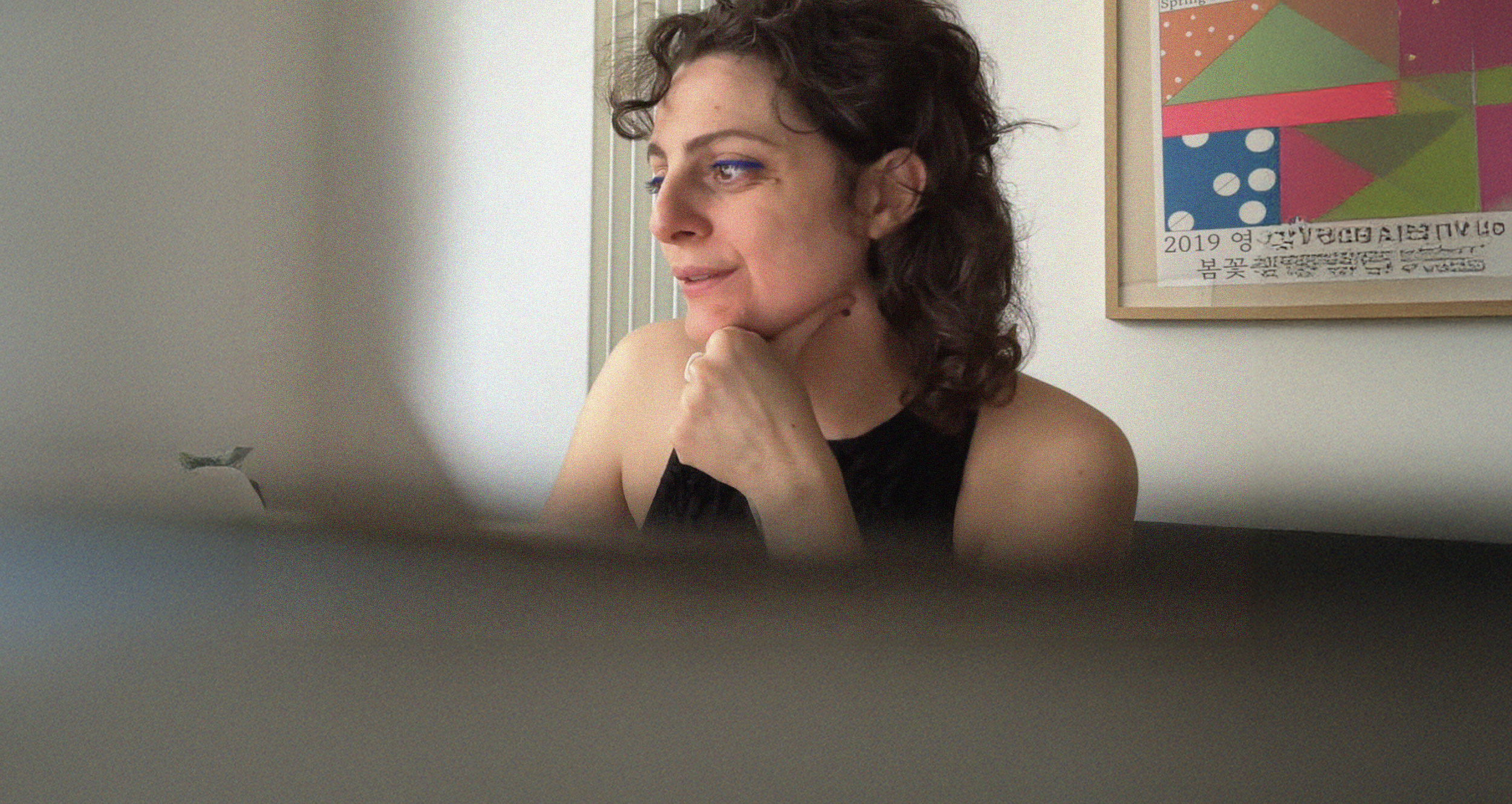 A woman with dark curly hair and blue eyes is sitting indoors, resting her chin on her hand, looking to the side. She is wearing a black sleeveless top. Behind her, there is a colorful geometric art piece on the wall with some text in Korean.